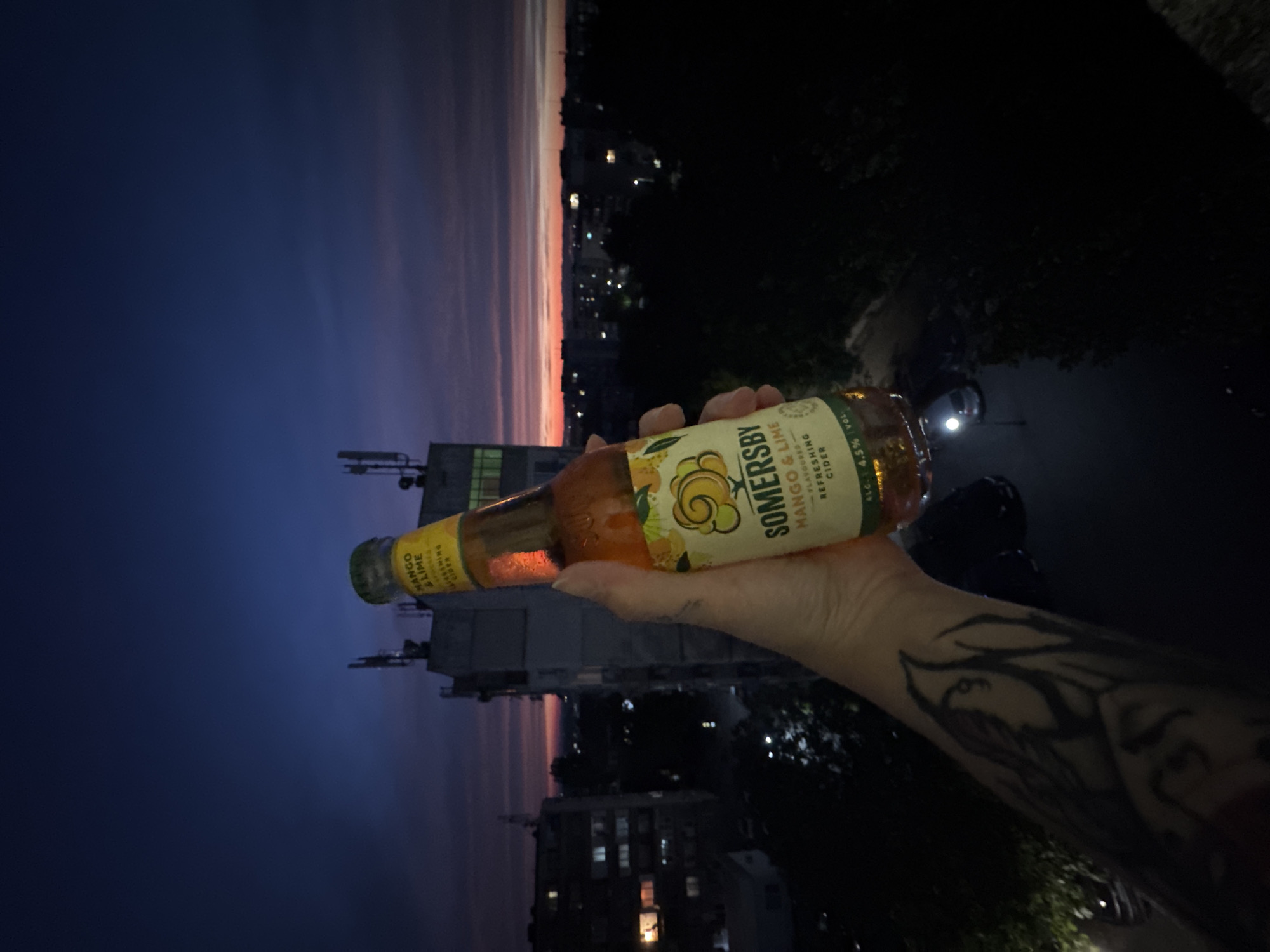 A hand, tattooed with dark imagery, holds a glass bottle of Somersby Mango & Lime flavored cider. The bottle is amber in color, with condensation visible on its surface. The label prominently displays the Somersby brand name and the flavor. The background depicts an urban landscape at dusk or dawn, with buildings and distant city lights visible against a gradient sky of deep blue and warm orange hues. The scene appears to be captured from a vantage point overlooking the city, possibly a balcony or rooftop.