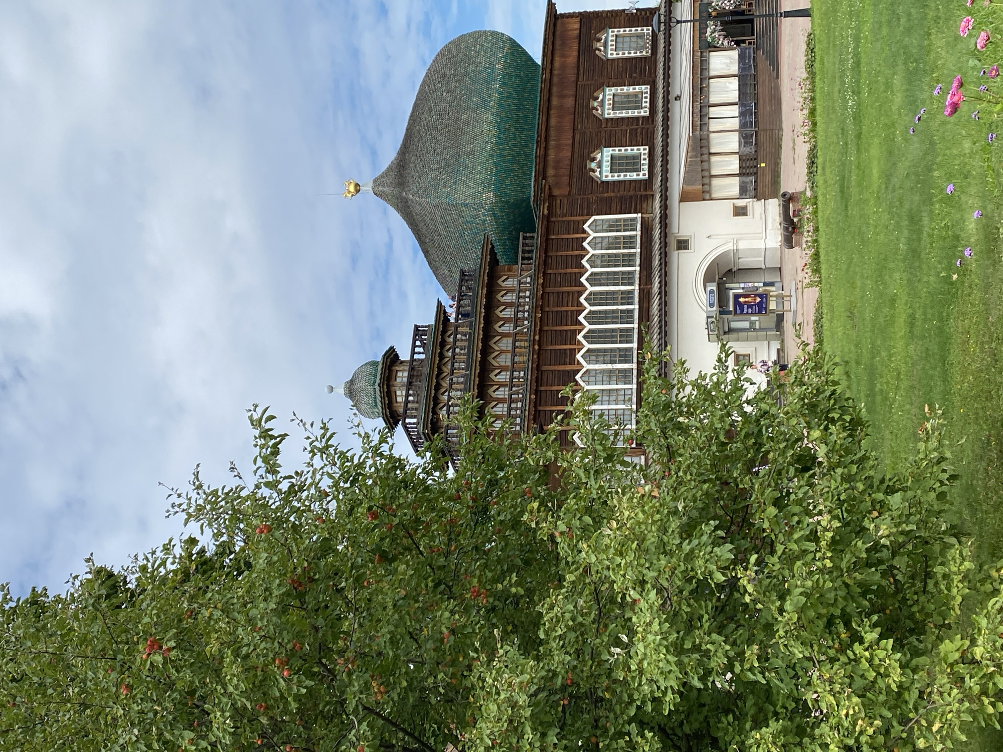 The image shows a view of a wooden building with a large, green, onion-shaped dome topped with a golden spire. The building has multiple levels with ornate wooden trim and decorative windows. In the foreground, a lush green lawn is visible, with some pink and purple flowers blooming. A large tree with red fruit is positioned to the left of the building, partially obscuring the view. In the background, the sky is cloudy with patches of blue.
This building is the unique wooden structure of the Izmaylovo Kremlin in Moscow, Russia. The Izmaylovo Kremlin is a cultural and entertainment complex built in a fantastical, fairy-tale style, inspired by medieval Russian architecture. It is located in the Izmaylovo District of Moscow.