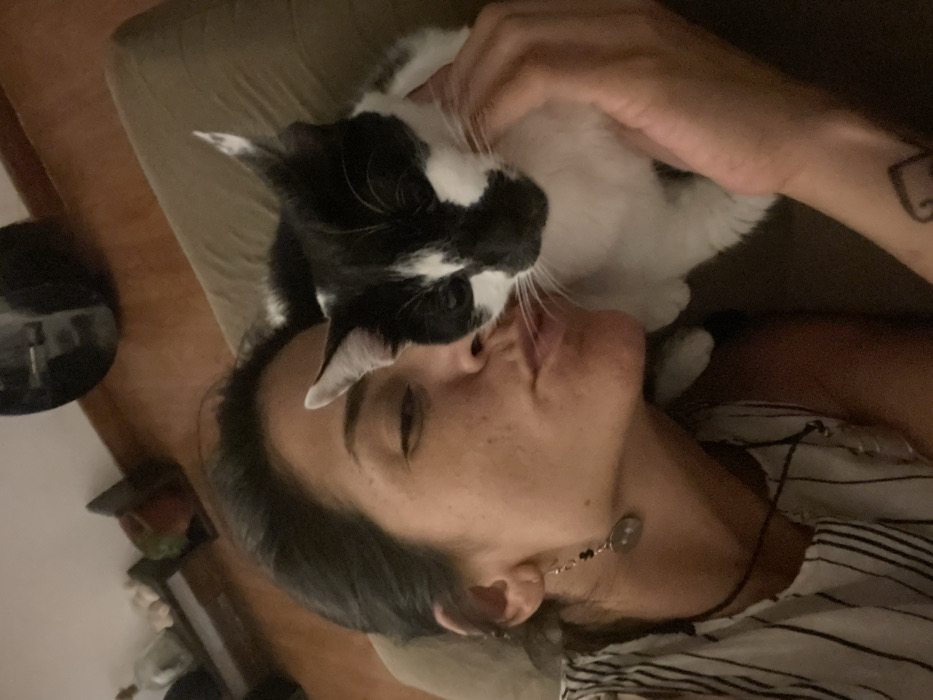 A woman with dark hair lies on a tan couch while a black and white cat sits on her face. The cat's nose is close to the woman's mouth, almost appearing to kiss her. The woman is wearing an earring with a circular pendant and a black and white striped shirt. A heart tattoo is visible on the woman's arm. A side table with objects on it is in the background.