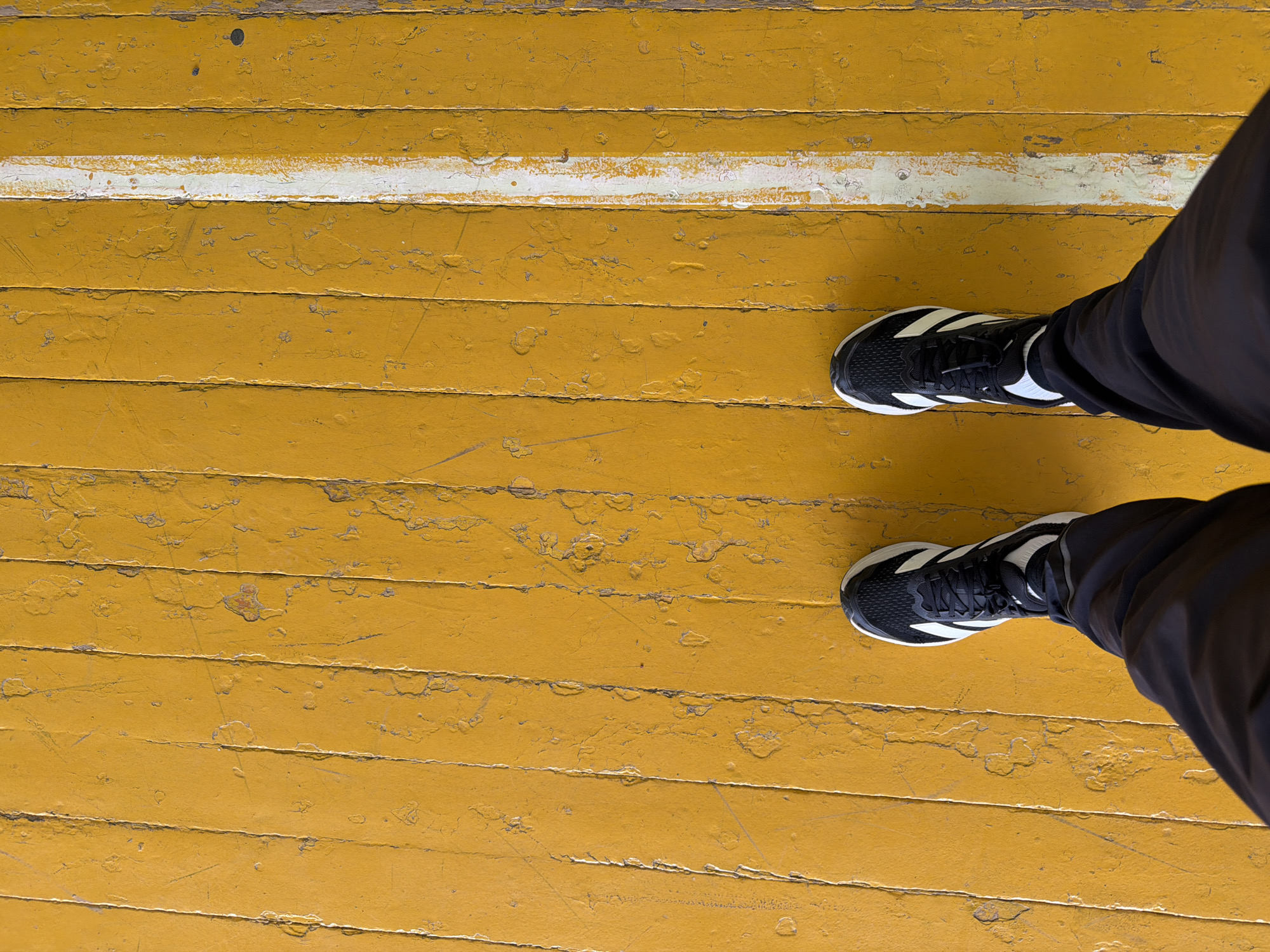 The image presents a top-down view of a person's feet and lower legs standing on a worn, yellow wooden floor. The floor is composed of horizontal wooden planks, painted a bright yellow, with significant signs of wear including peeling paint, chips, scratches, and areas where the underlying wood is exposed. A distinct, but also worn and chipped, white line runs horizontally across the upper middle section of the floor, spanning the width of the frame.
The subject is wearing black athletic shoes with prominent white stripes along the sides, and dark, possibly black, trousers or sweatpants. Both feet are placed parallel to each other, toes pointing towards the top of the frame, suggesting the person is standing still. The focus is primarily on the textured floor and the footwear.
There are no specific activities taking place other than standing. The lighting appears even, but the time of day or specific weather conditions are not discernible. The setting appears to be an aged indoor space or an outdoor area with a painted wooden surface, possibly a sports court or a weathered walkway in Izhevsk, Russia. No visible text is present in the image.
