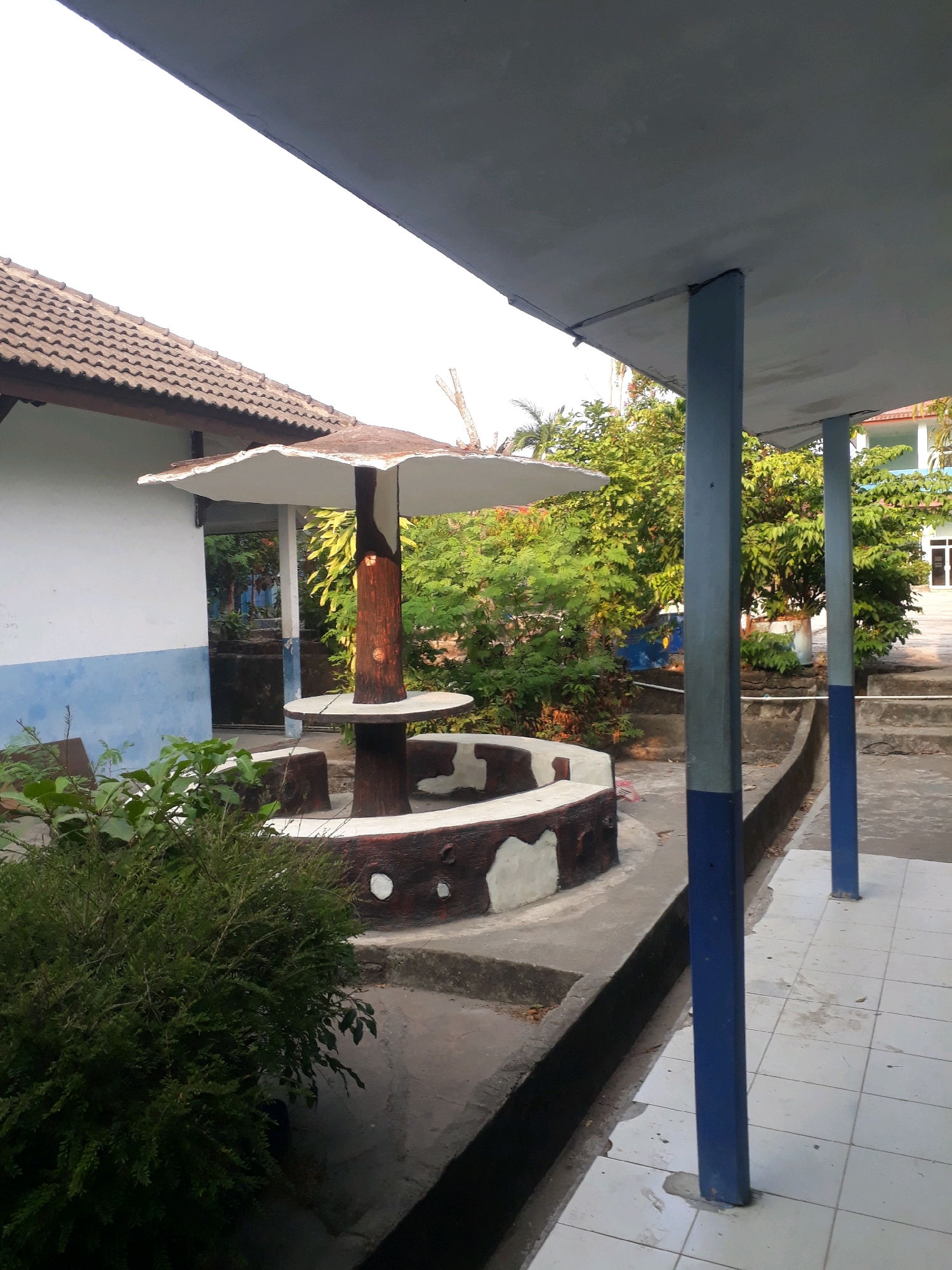An outdoor shot of a sheltered seating area with a mushroom-shaped canopy and a circular bench is captured. The seating area appears to be located in a public space, possibly a schoolyard or park, given the presence of a white building with a tiled roof and blue trim in the background, and lush green trees surrounding the area. The ground is paved with light-colored tiles, and a drainage ditch runs alongside the tiled walkway. Two blue pillars support the overhang of the structure. The image is taken from a low angle, with the sky and the overhang taking up a significant portion of the frame. No people are visible in the scene. The overall atmosphere is serene and tranquil. The image is an outdoor photograph from the city of Pangkalpinang, Indonesia.