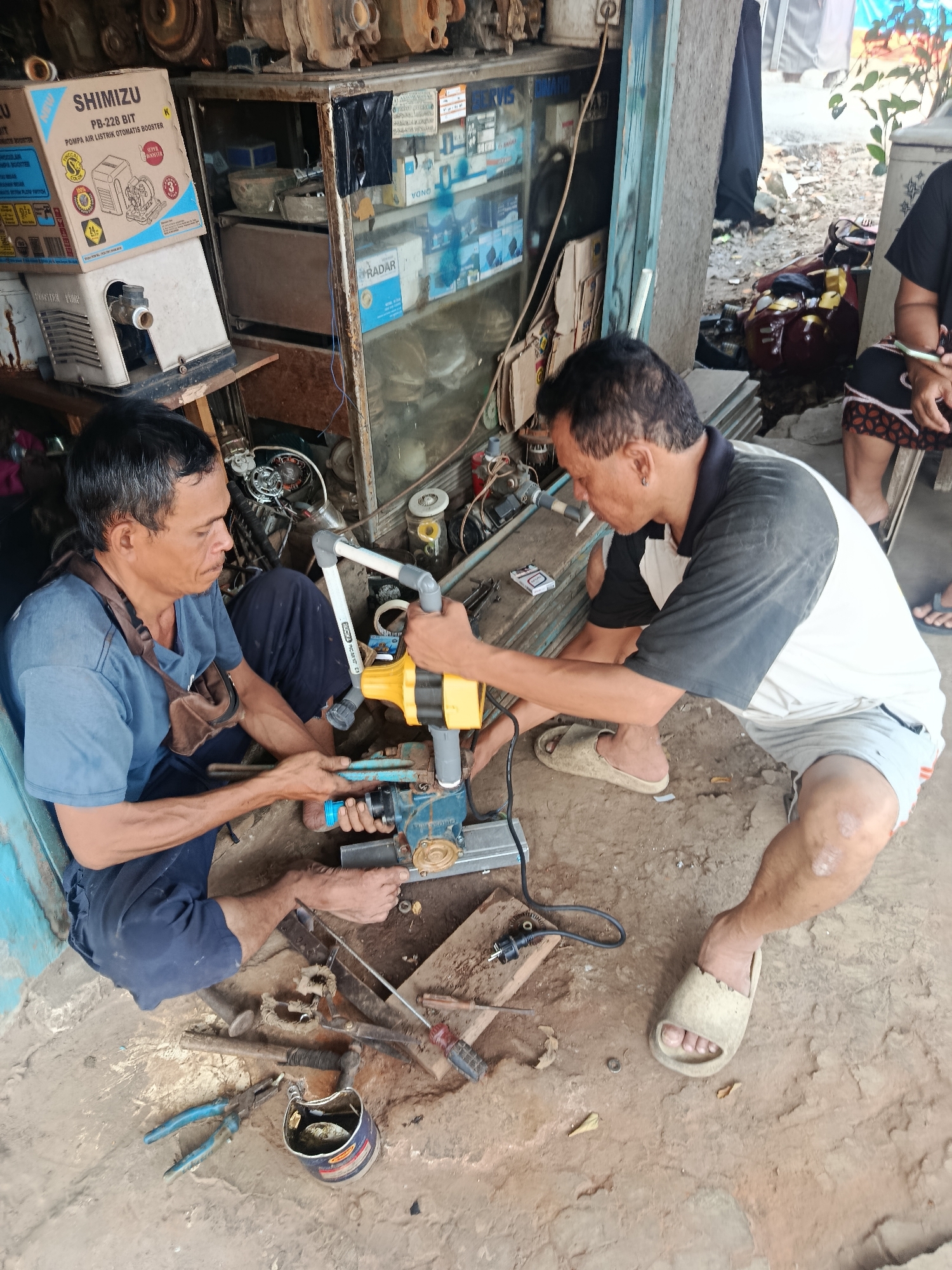 Two adult males are engaged in repairing a water pump at an outdoor workshop in Sampora, Indonesia. The man on the left, wearing a blue t-shirt and dark blue pants, is holding a pair of pliers and appears to be working on the pump's base. The man on the right, wearing a black and white t-shirt and grey shorts, is holding the yellow portion of the water pump and working on its connections. Tools and equipment are scattered on the ground, including various pipes, wires, a screwdriver, and a can of oil. In the background, a glass-fronted cabinet displays more water pump parts and boxes. The ground is made of packed dirt, and the overall environment suggests a busy, functional repair shop.