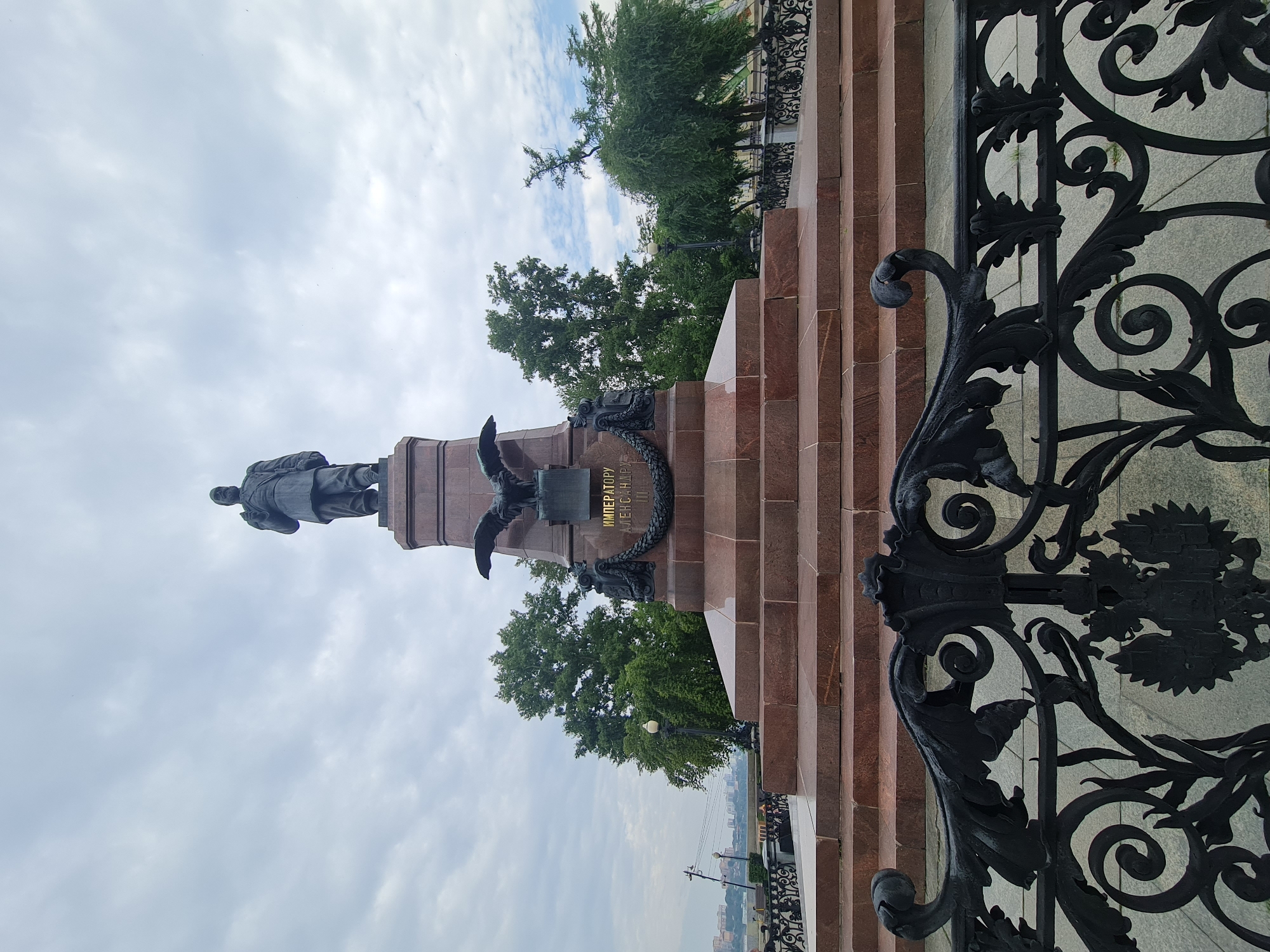 The image shows a monument in Irkutsk, Russia, dedicated to Emperor Alexander III. The monument is located on the embankment of the Angara River. In the foreground, a black wrought-iron fence with ornate scrolls and an emblem of the double-headed eagle can be seen. Behind the fence, a large, multi-tiered granite pedestal rises, topped by a bronze statue of Emperor Alexander III. The statue depicts him standing in a military uniform, holding a sword. On the pedestal, there are inscriptions and a wreath. The monument is surrounded by trees and on the horizon, the opposite bank of the river with buildings is visible. The sky is cloudy.