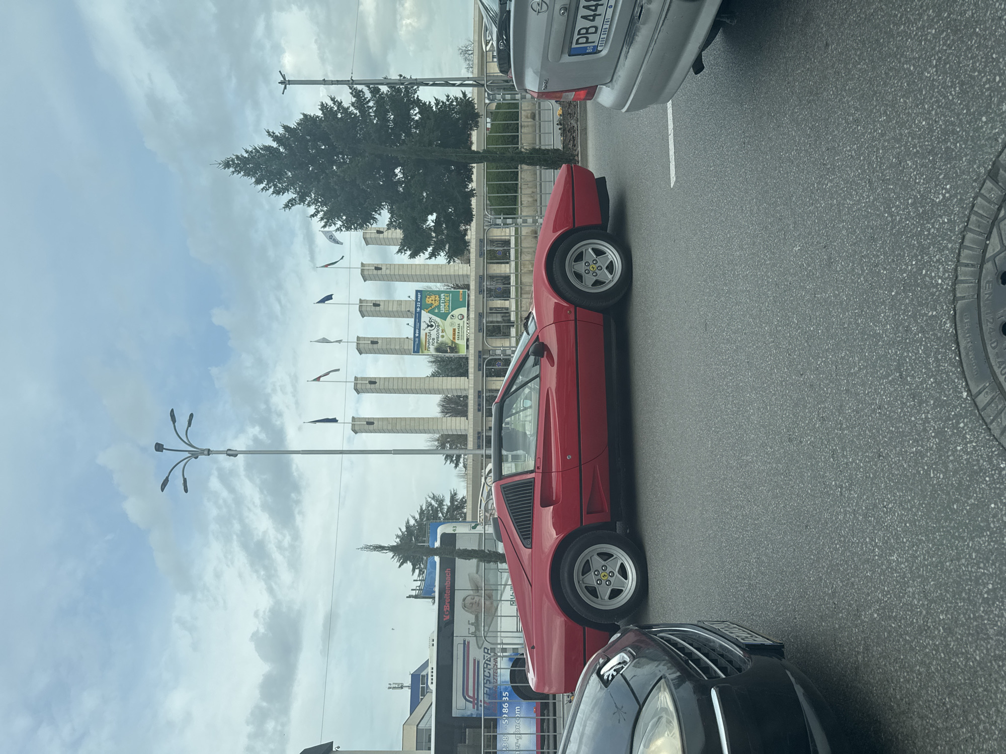 The image depicts a red Ferrari 308 GTS model stopped in traffic on a city street. There is a silver Opel and a black BMW around the vehicle. On the opposite side of the street is a tree line and a series of white columned structures. Banners or flags hang from each. A large advertising banner on one of the columned structures is visible. A street light is in the upper left. Several storefronts and business signs are visible. Street advertisements are to the right side of the column structure. The license plate of the Opel is visible. According to the license plate and the advertising, the location appears to be Plovdiv, Bulgaria.