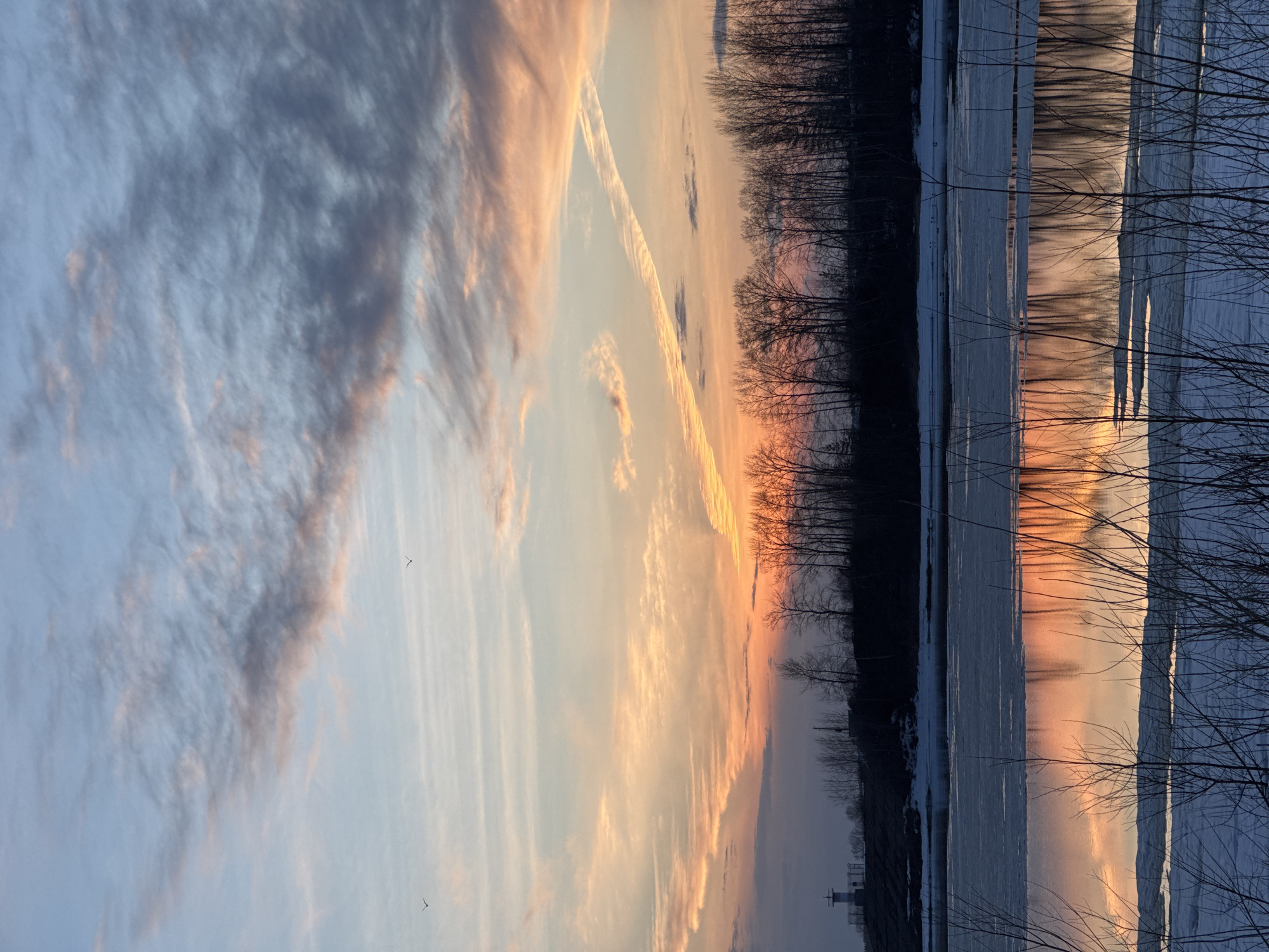 Here is a description of the image:
The image shows a sunset over a body of water in Chaikovsky, Russia. The sky is filled with clouds tinged with orange and pink from the setting sun. A contrail stretches across the sky. Silhouetted trees line the horizon. The water reflects the colors of the sunset, with patches of ice visible on the surface. In the foreground, bare branches of trees frame the scene. A structure, likely a lighthouse, is visible on the shore in the bottom left corner.