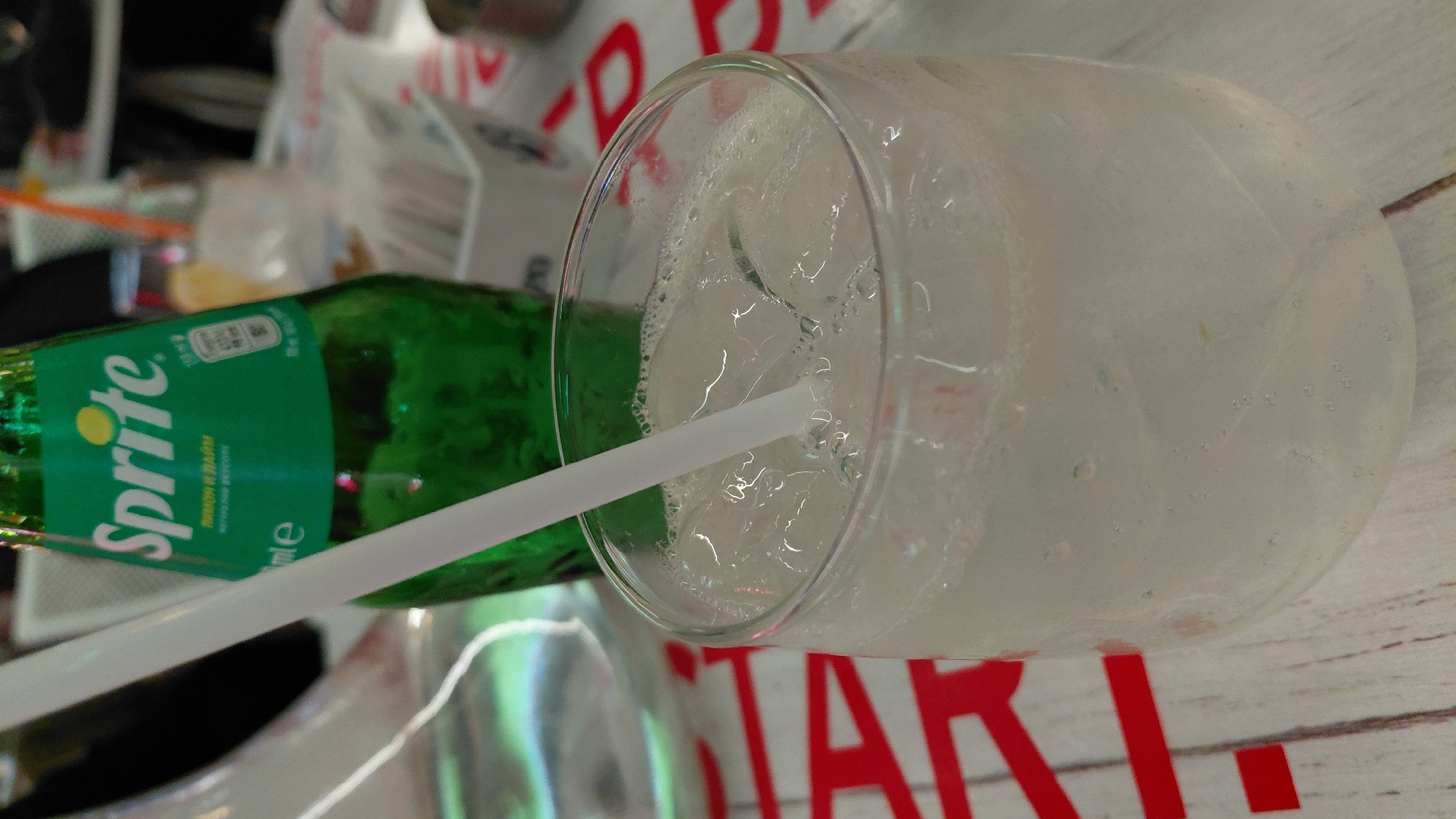 This close-up, diagonally oriented image captures a refreshing beverage setup indoors. The main subjects are a dark green glass bottle of Sprite, a clear glass filled with ice and a light-colored, fizzy drink, and a white plastic straw.
The Sprite bottle is prominently displayed on the left, partially obscured by the straw. Its label features the iconic white "Sprite" logo with a yellow dot, and visible text in Cyrillic script reads "Лимон и Лайм" (Lemon and Lime). Nutritional information, including "100ml" and a crossed-out "100" (likely indicating sugar content), is also visible on the label.
To the right, a clear, rounded glass holds a cold, bubbly drink with several ice cubes. The outside of the glass appears frosted or covered in condensation, suggesting the drink is very cold. A white straw is inserted into the drink, angled downwards into the liquid.
The activity depicted is the preparation or serving of a cold soft drink. The bottle is open, and the contents are in the glass, ready for consumption.
Notable details in the background are blurred but suggest an indoor setting. A light-colored wooden table surface runs horizontally across the bottom of the frame. Behind the bottle, out of focus, are elements that appear to be a white plastic bag or container, and partially visible red letters that could spell "STAR" or similar. No people are visible, and thus no emotions are conveyed. The time of day is not discernible.