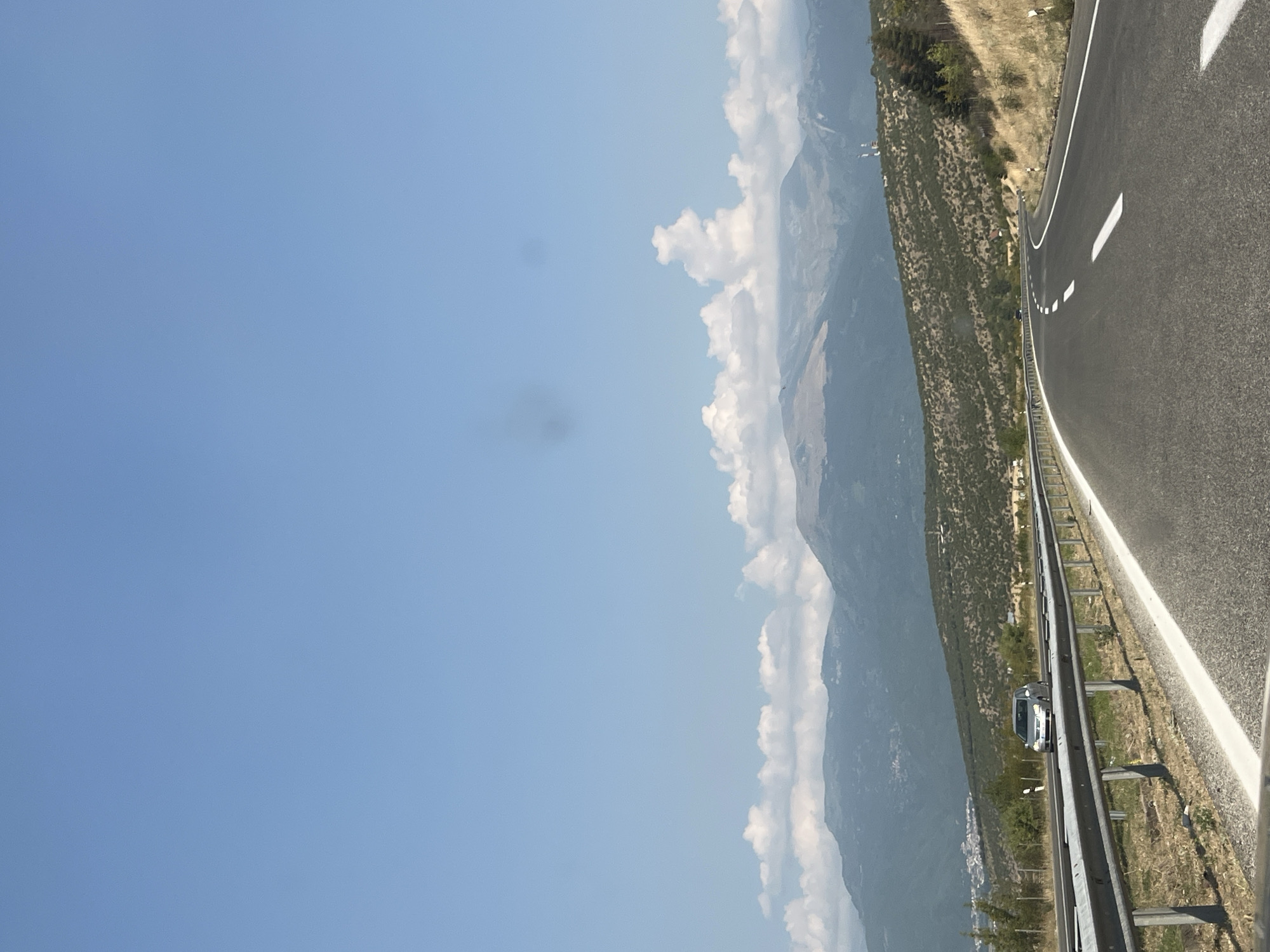 An exterior, vertical, medium shot captures a highway in Zhelino, North Macedonia, under a clear blue sky with scattered white clouds. The road, asphalt with white lane markings, curves gently to the right. A silver car is visible in the left lane, traveling away from the camera. A metal guardrail borders the left side of the highway, separating it from a dry, grassy embankment. In the middle ground, rolling hills covered in sparse, green vegetation rise towards distant, hazy mountains. Some buildings and trees are visible on the left side of the frame near the base of the hills. The overall scene depicts a daytime road trip through a mountainous, rural landscape.