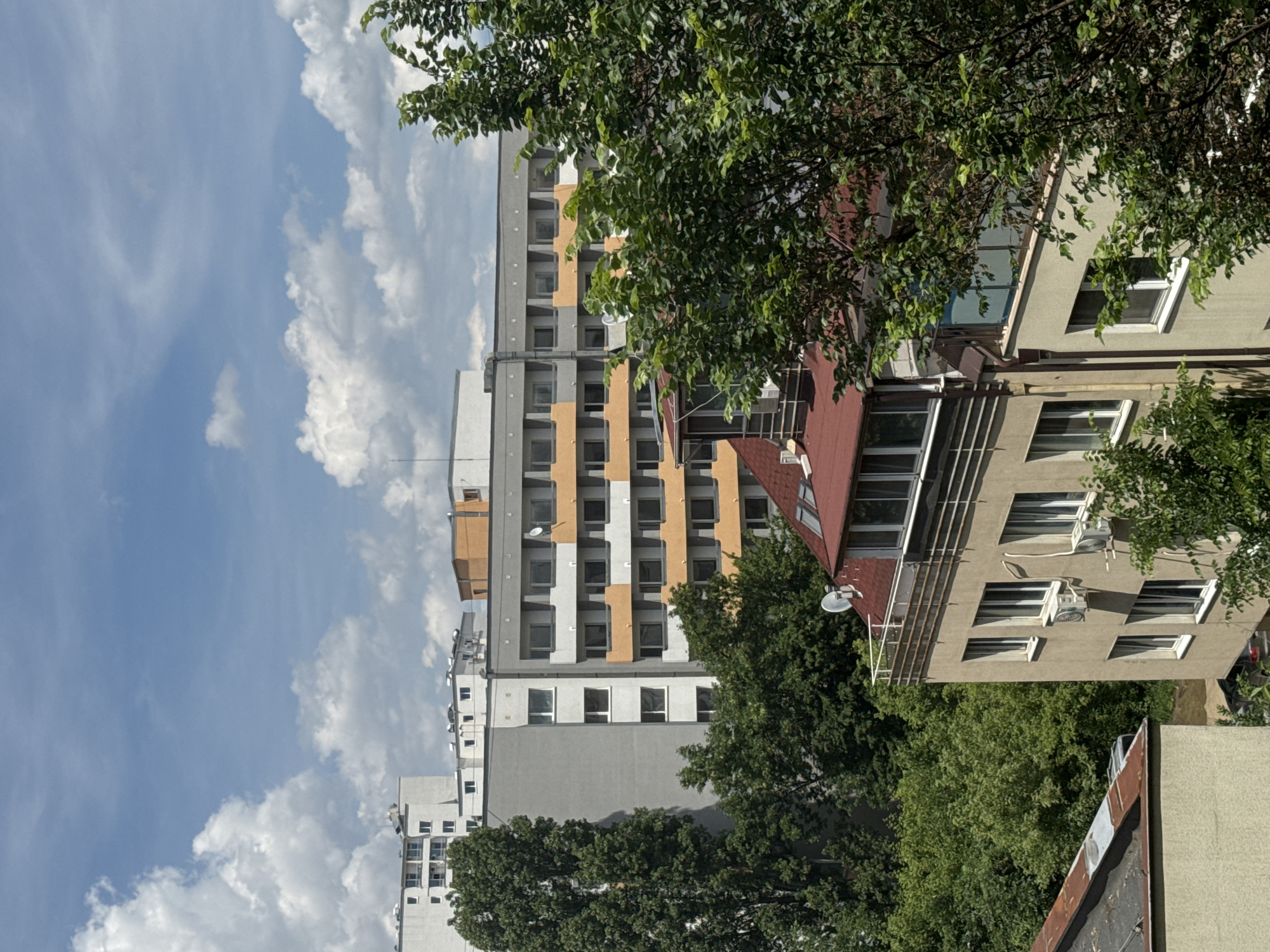 This is a medium-distance, eye-level, outdoor, daytime, vertical shot taken from an elevated vantage point, looking out over a cityscape. The image is dominated by a mixture of apartment buildings and lush green trees, set against a bright blue sky with scattered white cumulus clouds.
In the foreground, on the lower right, a beige apartment building with a brown roof is partially visible. It features several windows with white frames, and two air conditioning units are mounted on its facade. To the left of this building, another building with a weathered, rusty metal roof is seen from a low angle.
In the mid-ground, a larger, light gray apartment building with multiple floors is prominent. This building has a repeating pattern of balconies with orange accents and dark window frames. To its left, a very tall, slender tree with dense green foliage stands out. Further back, another apartment building is visible, this one primarily white with a darker horizontal band and multiple windows.
In the background, more apartment buildings of varying heights and architectural styles can be seen, stretching towards the horizon. Some appear to be older Soviet-era panel buildings, while others have more modern designs. The sky is a vibrant blue, with large, fluffy white clouds casting soft shadows on the buildings below. The overall impression is a dense urban environment interspersed with abundant greenery, suggesting a residential area within a city. The architecture and building styles are typical of Eastern European cities, and the presence of specific building types, such as the characteristic panel blocks, aligns with urban landscapes found in Sofia, Bulgaria.