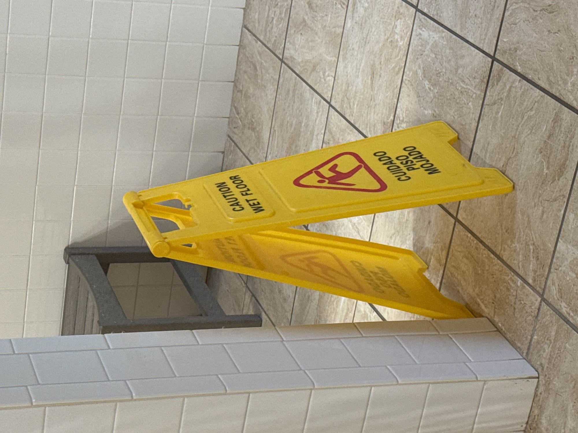 A yellow "Caution Wet Floor" sign is placed in a bathroom. The sign has a graphic of a person slipping and the text "CUIDADO PISO MOJADO" in Spanish. The floor is tiled with a light brown pattern. The base of the sign rests on a tiled surface. To the left of the sign is a dark grey object. The image was taken in Myrtle Beach, United States of America.