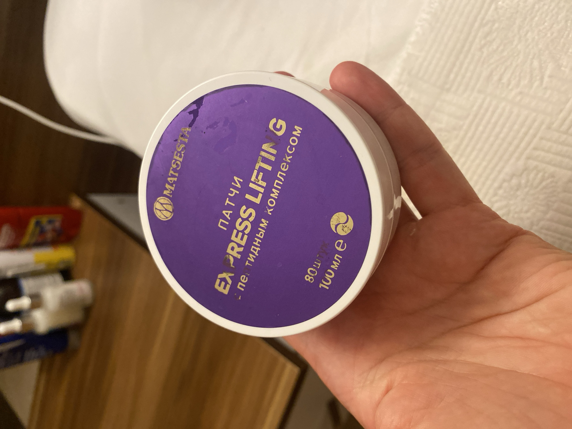 A hand holds a round, purple cosmetic container with white lettering and a white rim. The container has a gold logo with the text "MATSESTA" above the words "PATCHI EXPRESS LIFTING". Below this, in smaller text, it reads "- пептидным комплексом". At the bottom of the container, there is text in Russian and a gold circular symbol. The background is blurred and appears to be a wooden surface with some bottles on it, and to the right, a white textured surface. The image was taken in Aksu, Turkey.