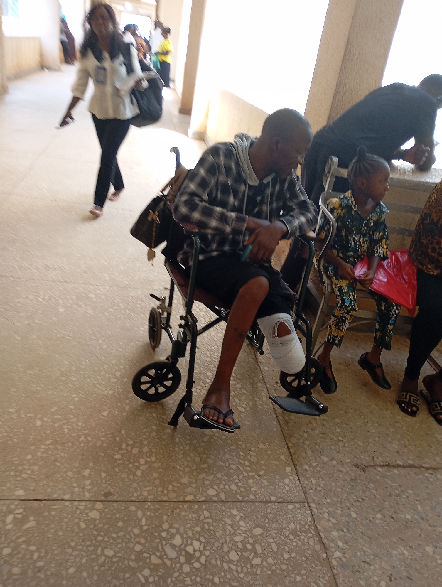 The image shows a man sitting in a wheelchair with a bandaged lower leg. He is wearing a plaid hooded shirt and shorts, and sandals. He is holding a smartphone. A young girl is sitting next to him on a chair, holding a red plastic bag. A woman is standing near the man and the girl. In the background, a woman is walking in the hallway with a purse slung over her shoulder. The location is a hallway with beige floors and walls. The location is the city of Gwong, Nigeria.