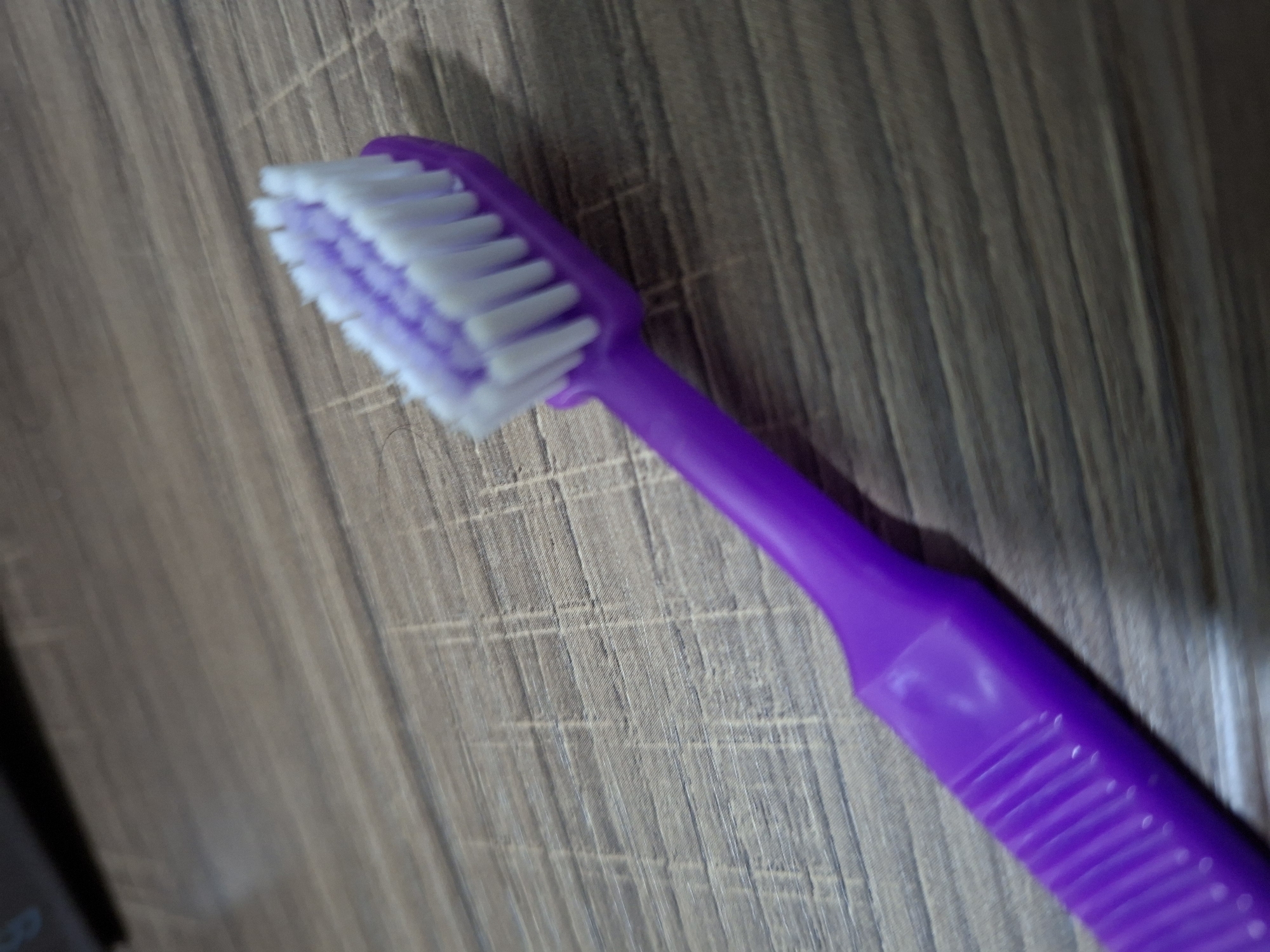 A purple toothbrush with white and purple bristles lies diagonally on a wooden surface. The toothbrush is in focus and positioned from the bottom left to the upper right of the frame. The wooden surface has a textured, horizontal grain pattern. The lighting appears to be natural, casting subtle shadows. There are no people or other objects visible in the image. The scene depicts a close-up view of the toothbrush and its immediate surroundings. The location in Samarinda, Indonesia is not discernible from the image.