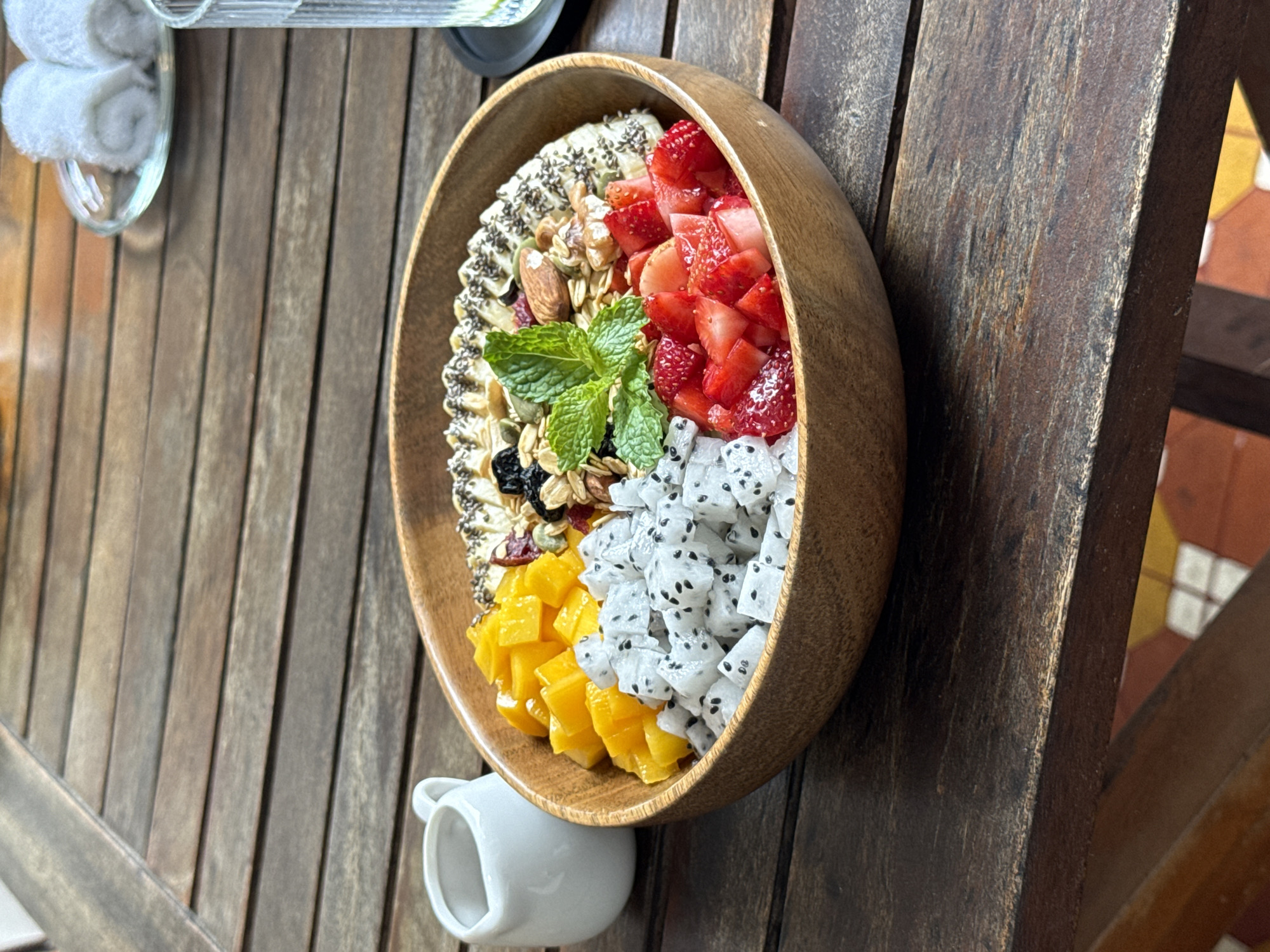 On a weathered, wooden table sits a partially filled wooden bowl. The bowl contains four distinct sections: sliced bananas topped with chia seeds, a mix of granola, nuts, and dried fruit, diced strawberries, and diced dragon fruit. A sprig of mint garnishes the center. A small, white, ceramic creamer is positioned beneath the bowl, supporting it. In the upper left corner, a glass containing tightly rolled white towels is visible. The scene is likely set outdoors at a cafe or restaurant, evidenced by the wooden table and the style of presentation. Although it cannot be determined from this image, it may be located in the city of Đà Nẵng, Vietnam.