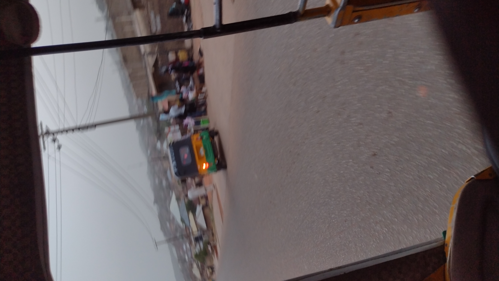 The image depicts a street scene, likely in a densely populated area within Jos, Nigeria. The perspective is from inside a moving vehicle, possibly a tuk-tuk or auto-rickshaw, with the road filling the majority of the frame. Another vehicle, possibly another tuk-tuk, is visible ahead on the same road. There are indistinct human figures on the roadside, suggesting market stalls or pedestrian activity. Utility poles with power lines are visible along the left side of the road. The overall light suggests either early morning or late afternoon.