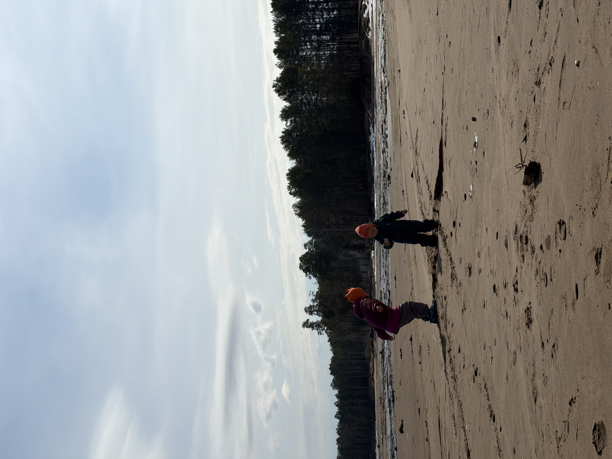 The image shows two children on a sandy beach. A forest lines the back of the beach. The sky is cloudy. The location is likely near Chaikovsky, Russia, based on the image's origin. Both children are wearing orange hats. One child is wearing dark clothing, the other child is wearing dark clothing and pink pants. The children appear to be playing on the beach. The sandy beach shows numerous footprints.