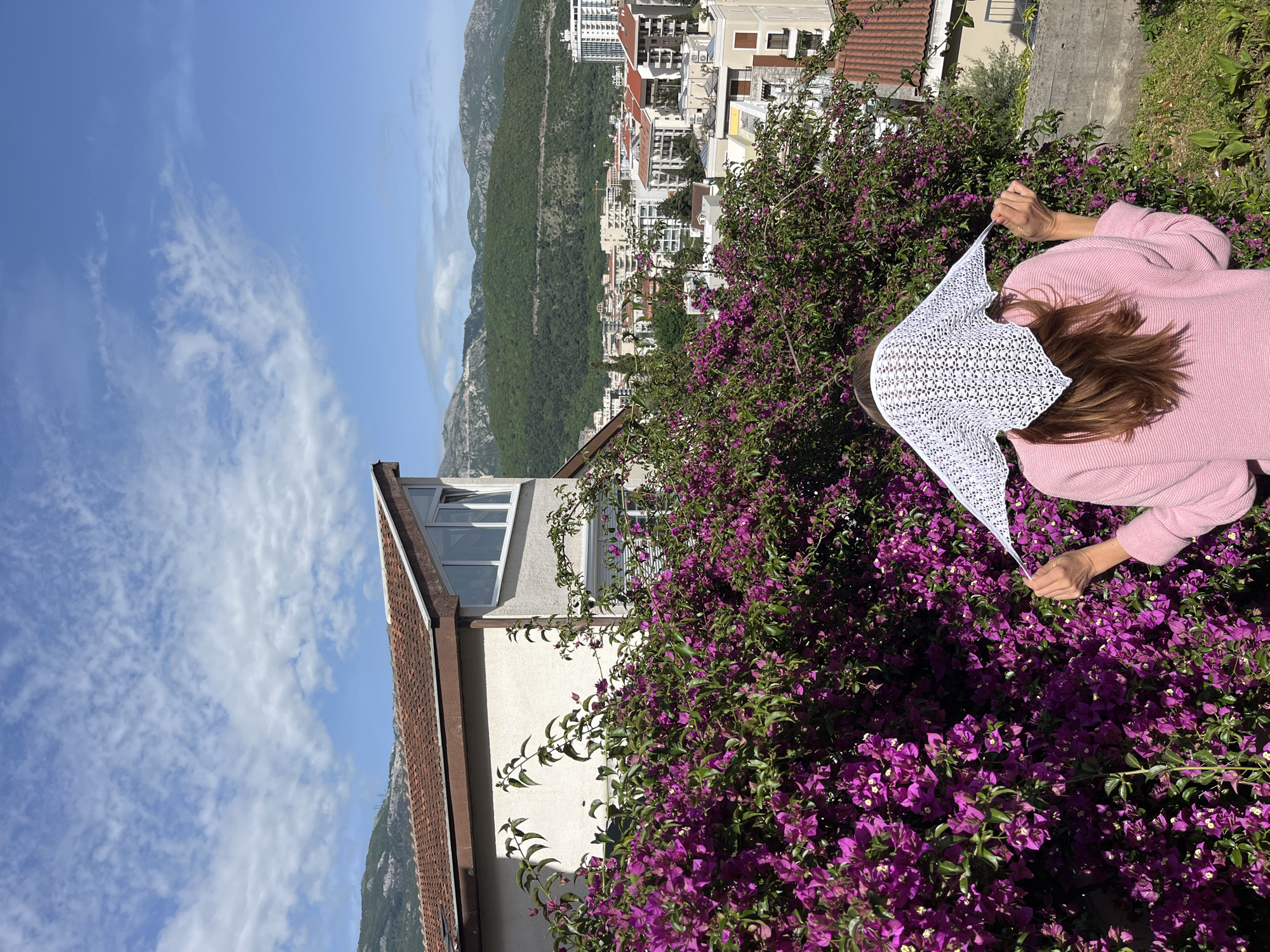 A medium shot of a woman wearing a pink sweater, positioned in the right side of the frame and partially obscured by a large bougainvillea bush with vibrant purple flowers. She is holding a white lace head covering. In the background, the city of Budva, Montenegro, is visible, including buildings, mountains, and a clear sky with scattered clouds. The perspective is slightly tilted. A building with a red-tiled roof is to the left side, and a mountain range extends along the skyline.