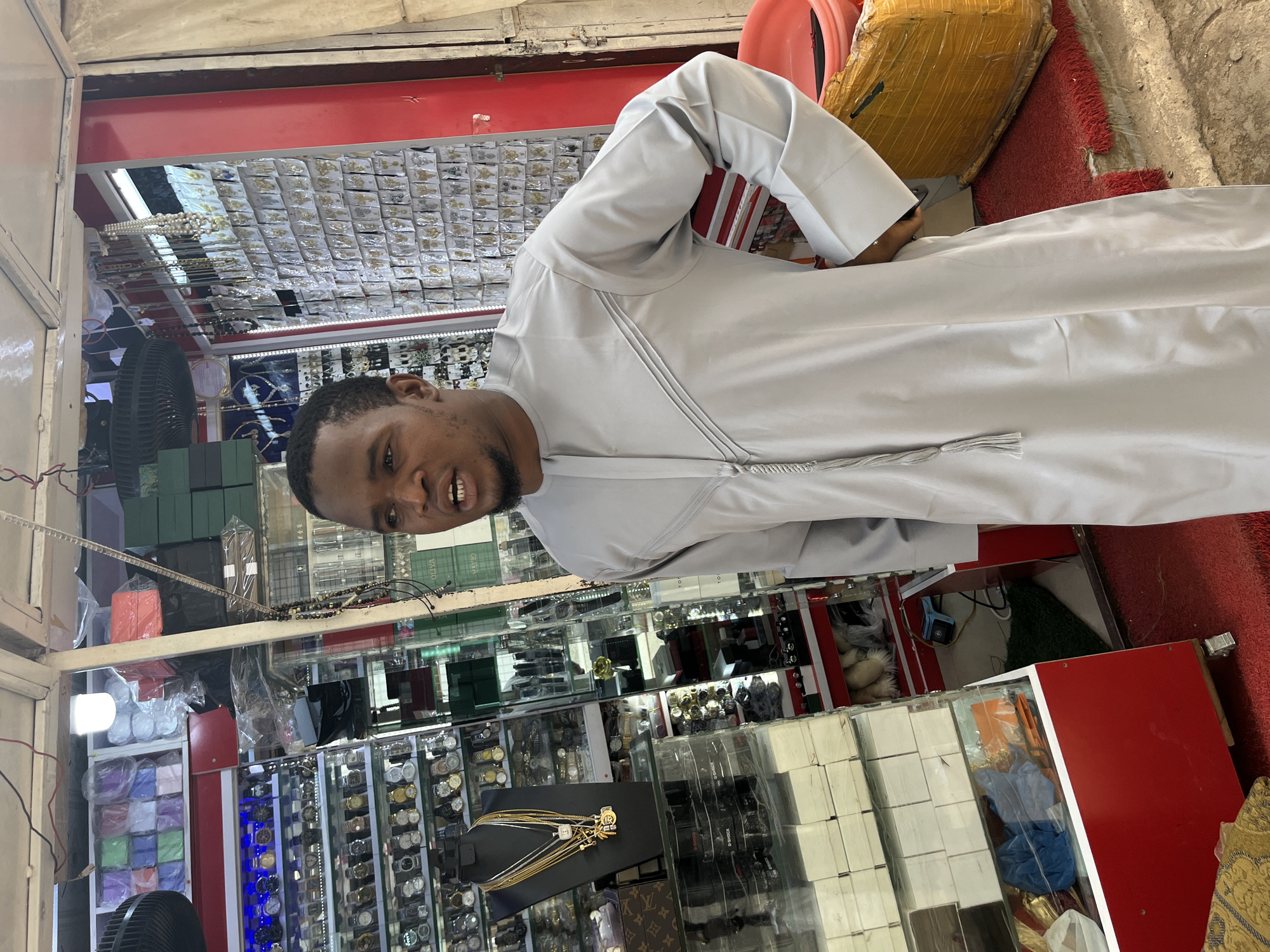 A man stands in a shop in Abuja, Nigeria, wearing a light gray kaftan with decorative stitching and a tassel. The shop appears to be a retail establishment selling accessories, with displays of watches, jewelry, and other assorted items visible in the background. The man is looking to his right, and his mouth is open as if speaking. The shop's interior is brightly lit, and the overall impression is of a busy marketplace environment.