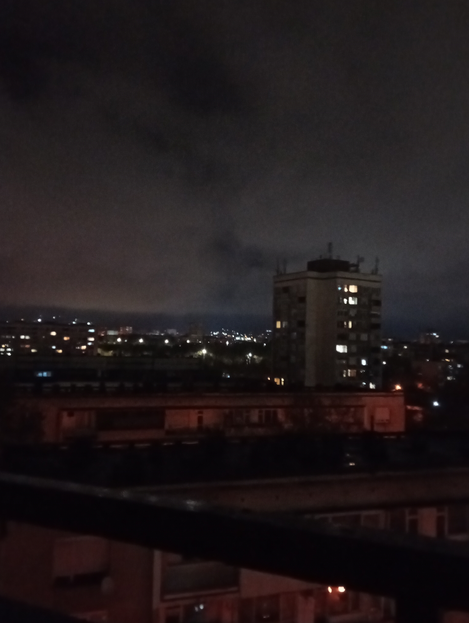 This image presents a nighttime view of an urban landscape, likely captured from an elevated position such as a balcony, suggested by the dark railing in the foreground. The overarching impression is one of deep night, with a dark, overcast sky dominated by heavy clouds that obscure any stars or moon.
In the mid-ground, a prominent, multi-story, rectangular building stands as a central subject. This structure, possibly an apartment block or office building, is partially illuminated by numerous lights emanating from its windows, indicating active occupancy. Antennas or similar fixtures are visible on its roof. Below and around this main building, other lower-rise structures are dimly discernible, some also displaying illuminated windows.
Beyond these immediate structures, the cityscape extends into the distance, marked by a widespread scattering of artificial lights. These lights, likely from various buildings, streets, and other urban sources, create a luminous horizon that contrasts with the dark sky and terrain.
The scene is devoid of any visible people or discernible activities, offering a quiet, static panorama of the city after dark. The weather conditions appear to be cloudy, contributing to the muted and somewhat somber atmosphere. There are no identifiable location cues or visible text within the image.