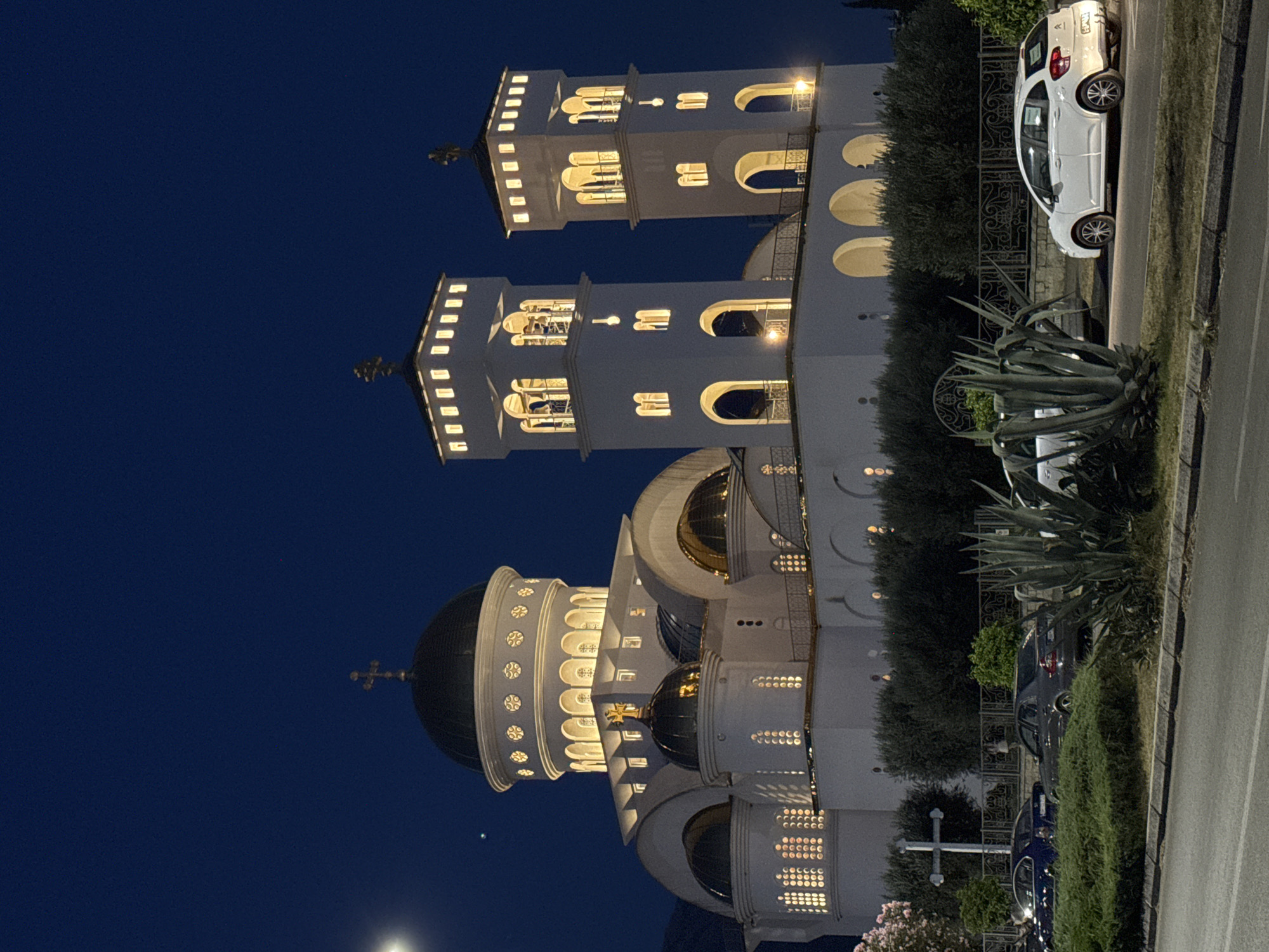 This media file depicts a church, likely an Orthodox cathedral, at night in Bar, Montenegro. The main subject is the illuminated white church building, featuring prominent domes and bell towers. Two tall, white bell towers with arched windows and decorative elements rise from the structure. The larger dome of the church is dark, contrasting with the brightly lit windows of the building. A cross is visible atop one of the towers and also a smaller cross on a smaller dome in front of the main dome.
In the foreground and along the right side of the image, a white car is parked on a street next to a grassy area and some bushes. A dark-colored car is also partially visible further down the street. The scene appears to be a street adjacent to the church, with a sidewalk or road edge and a well-maintained grassy verge with some plants, including large agave plants. The time of day is clearly night, indicated by the dark sky and the illumination from the church and streetlights. The lighting from the church casts a warm glow, creating a serene and somewhat majestic atmosphere. No people are visible in the image. There is a sticker on the back window of the white car, but no text is legible.