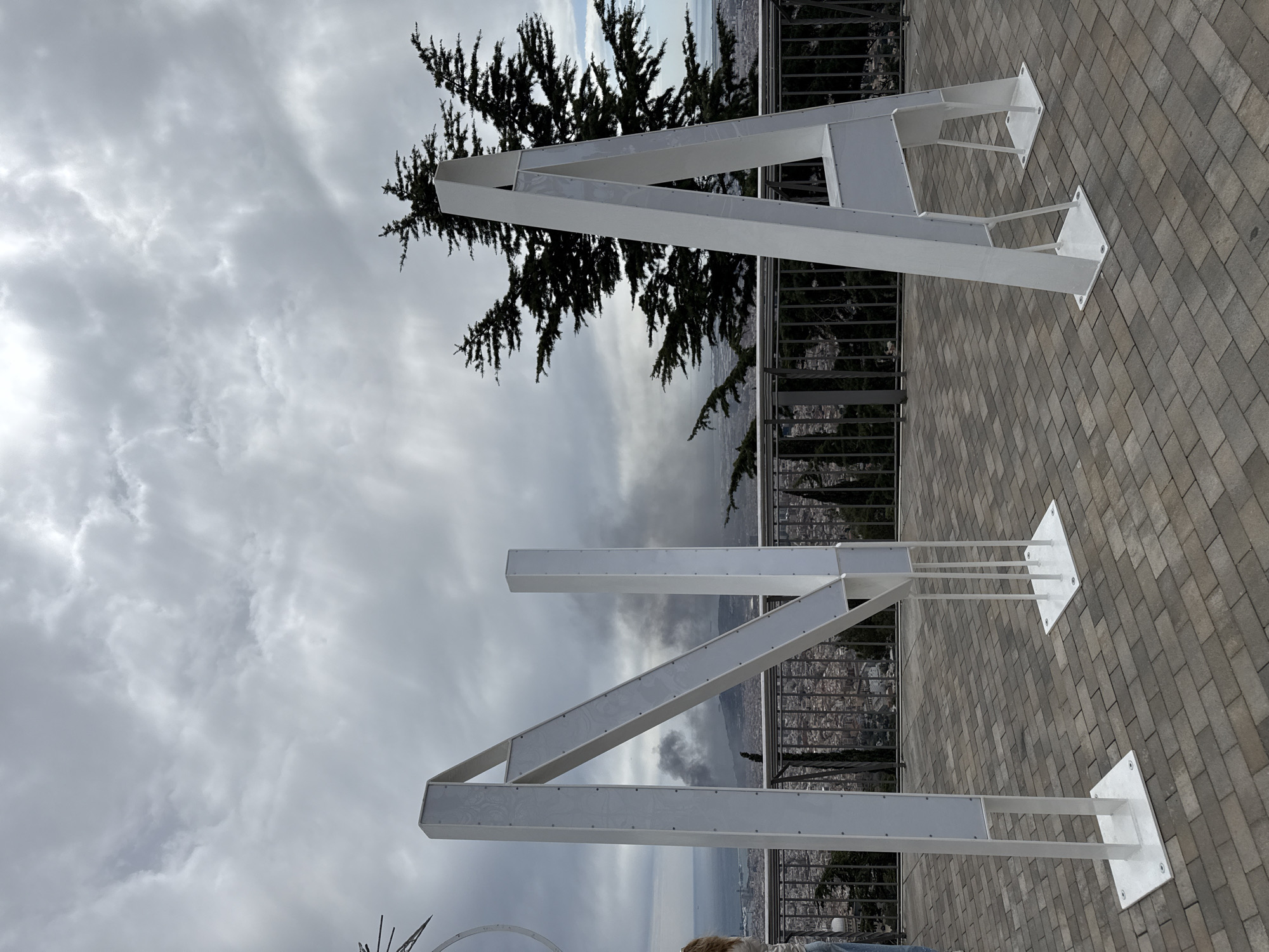 The image shows two large, white, metal letters, "A" and "N," presumably part of a larger sign or installation. The letters are situated on a gray brick-paved surface, likely a terrace or overlook, with a metal railing visible in the background. Beyond the railing, there's a view of a city with a cloudy sky overhead. A tree is located behind the letter "A". The letters appear to be supported by metal posts attached to square bases on the ground.
The location is implied to be Barcelona, Spain, based on the prompt. The view beyond the railing suggests a vantage point overlooking the city. No people are visible in the frame.
