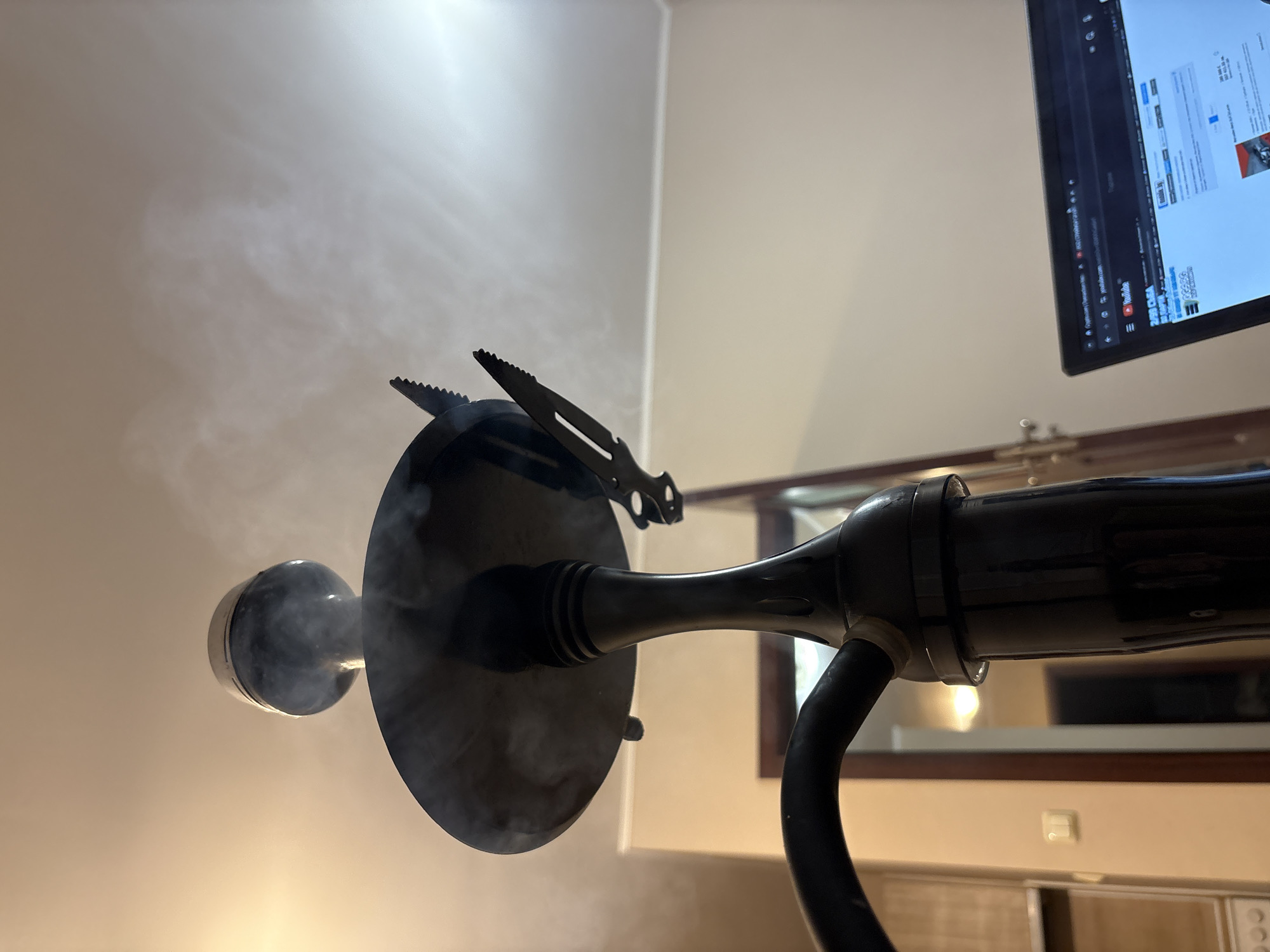The image shows a close-up of a lit hookah with smoke billowing around it. The hookah appears to be black metal, with a circular tray and a hose. A set of hookah tongs rests on the tray. In the background, a television screen displaying a YouTube page is visible. The room has neutral-colored walls, and a wooden door frame is partially visible. The lighting suggests an indoor environment. The specific location within Plovdiv, Bulgaria cannot be determined from the provided visual information.