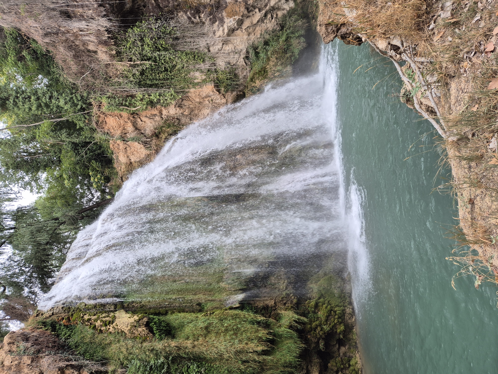 A wide waterfall cascades down a rocky cliff face into a tranquil, turquoise pool. Lush green vegetation clings to the sides of the cliff, interspersed with patches of reddish-brown rock. Trees with dense green foliage form a canopy overhead, suggesting a natural, secluded setting. The water is a vibrant shade of blue-green, with white foam where it crashes into the pool. The foreground shows a rocky bank with dry grass and fallen leaves. The overall impression is one of natural beauty and serenity. The location is Unknown, Unknown. There are no people or animals visible in the image.