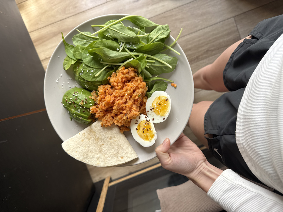 A first-person perspective photograph shows an individual holding a light gray round plate laden with food over their lap. The individual wears a white ribbed long-sleeved top and dark gray athletic shorts, partially revealing a white Nike logo. On the plate, arranged clockwise from the top left, are: a folded light beige tortilla, multiple slices of green avocado topped with small dark seeds, a mound of vibrant orange-red cooked lentils, a cluster of fresh green spinach leaves seasoned with coarse white salt, and two halves of a hard-boiled egg displaying bright orange-yellow yolks, also topped with small dark seeds. The background features light-colored wooden floorboards and a dark, possibly black, vertical surface on the left. A light-colored wooden furniture piece and a light-toned cushion are partially visible on the left. No specific visual cues are present to determine a precise location within the city of Tel Aviv-Yafo, Israel; the setting appears to be a generic indoor environment.