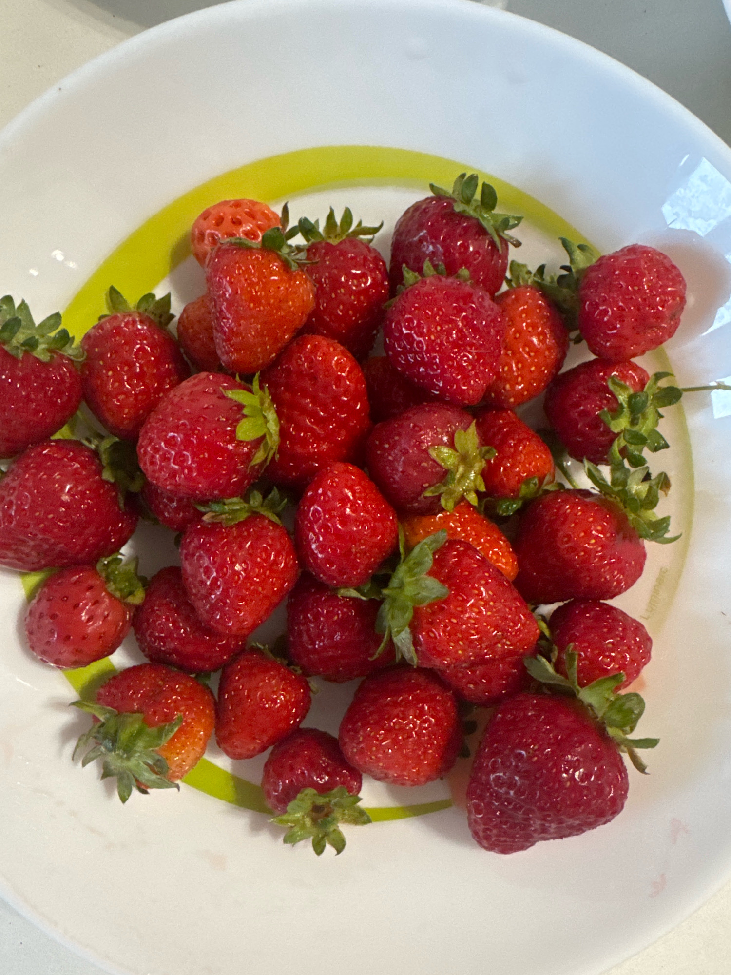 A close-up, overhead shot captures a white bowl filled with numerous fresh, ripe strawberries. The strawberries are predominantly a vibrant red, with some exhibiting lighter red or orange hues. A bright green stripe circles the rim of the bowl, visible along the top and right edges of the image. The strawberries are scattered unevenly, creating a visually textured arrangement. The lighting is bright, highlighting the glossy surfaces of the fruit. The scene is devoid of any people, actions, or interactions. The exact location in the city of Sovkhoza imeni Lenina, Russia, cannot be determined from the visual information provided in the image.