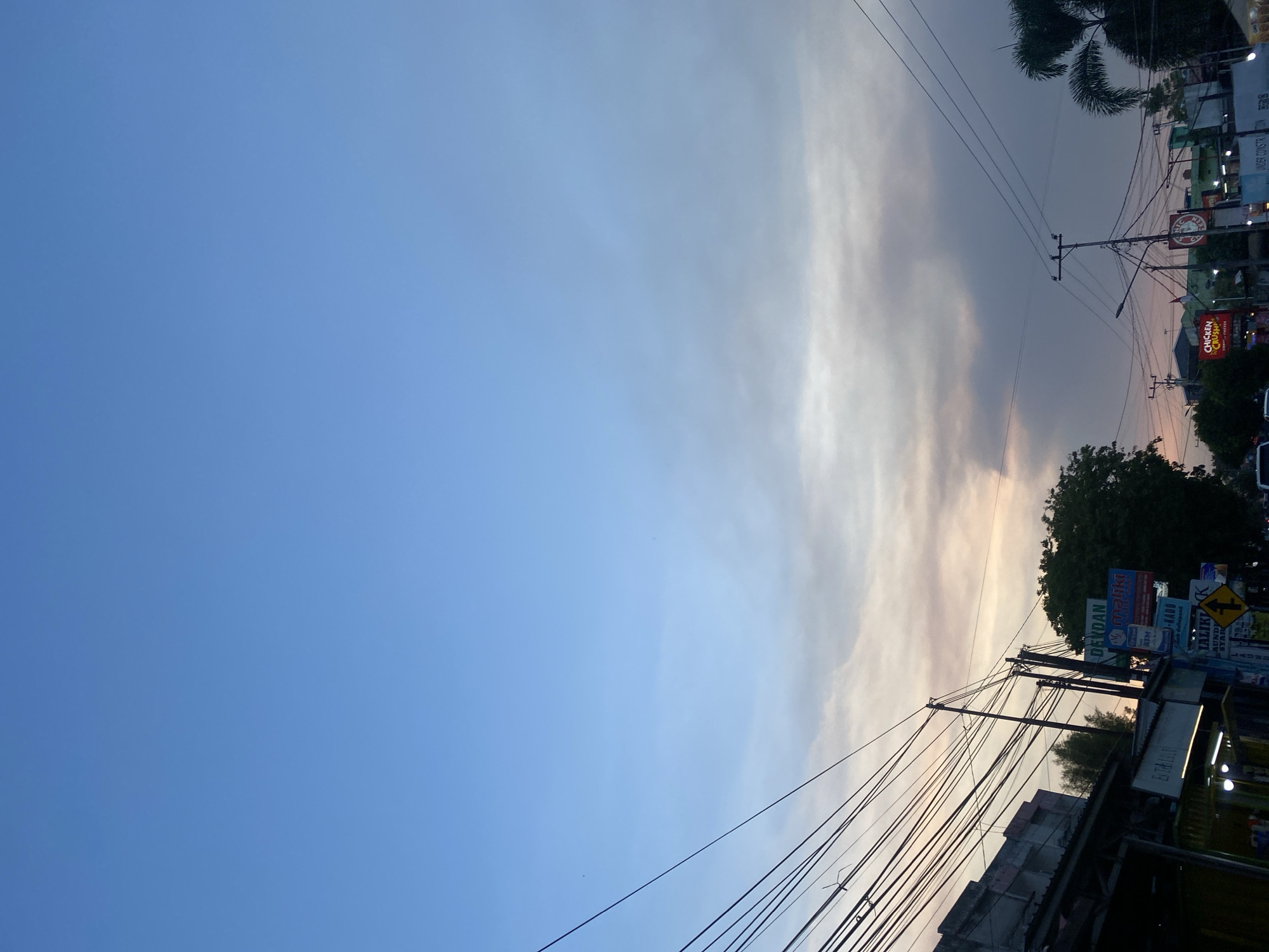 This is a low-angle, full shot taken at dusk on Jalan Trikora, Banjarbaru, Indonesia. The sky is a gradient of light blue and soft grey clouds with hints of pink from the setting sun. Thick power lines crisscross the upper left of the frame, angled diagonally from the left. On the left side of the road, there are buildings with signs, including one that reads "DEKORASI MALIKI" with an ATM machine. A large, dark green tree is visible behind the buildings. The right side of the image shows more power lines and utility poles. Further down the road, there are more buildings and signs, including one for "CHICKEN CRUSH" and a green and white flag. A palm tree is visible on the far right. The scene is illuminated by a mix of natural twilight and artificial light from the buildings. There are no people clearly visible in the image.