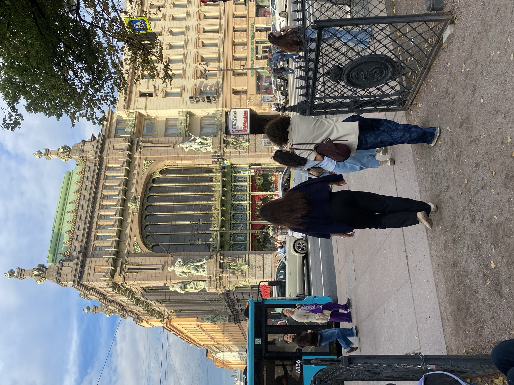 The image depicts the Singer Building, a historic Art Nouveau building in Saint Petersburg, Russia, located at Nevsky Prospekt, 28. The facade of the building is ornate, with a green roof and decorative elements. In the foreground, four women are walking on a sidewalk. The woman closest to the viewer is wearing a dark blue jacket and black pants, with her back to the camera. To her left, a woman in a light-colored trench coat and blue patterned pants walks ahead. Further to the left, two women walk side-by-side, one in a dark jacket and jeans, and the other in a light-colored cardigan and purple pants. A teal bus with the number "6856" on its side is visible on the street, along with several cars. The street is lined with buildings and a pedestrian crossing is marked. The sky is partly cloudy, and the overall lighting suggests it is either morning or late afternoon. A decorative black metal fence with a crest is on the right side of the image.