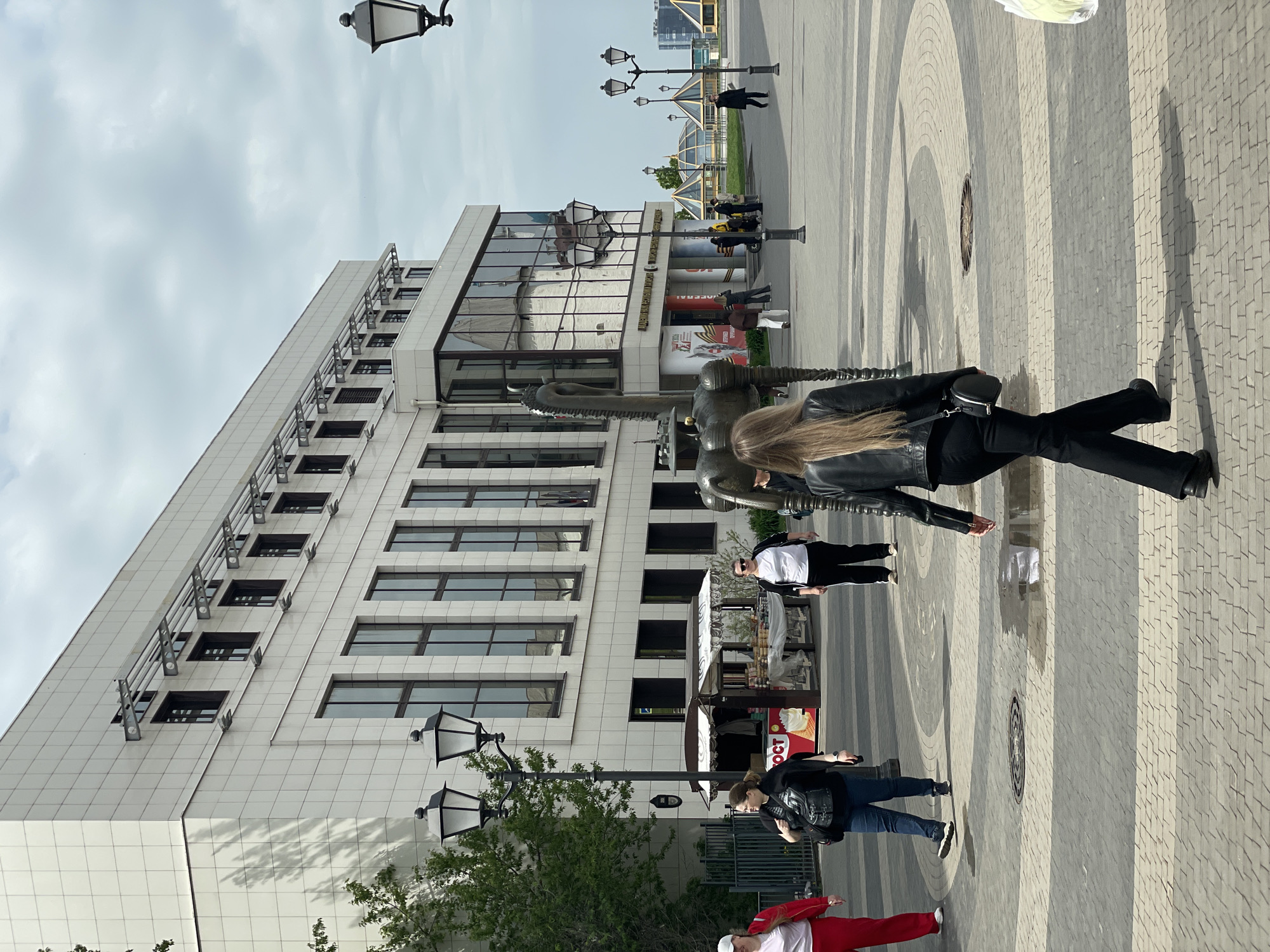 The image depicts a scene in Kazan, Russia, showcasing a building with numerous windows and a facade featuring a combination of white tiles and a reflective glass section. Several people are present; one woman in a black leather jacket and pants is prominently in the foreground. A bronze sculpture stands beside her. Other people stroll in the background, some near a kiosk with a white canopy. Street lamps are interspersed throughout the scene. The ground is paved with gray and white patterned tiles. The sky is overcast. In the distance, modern glass buildings can be seen.