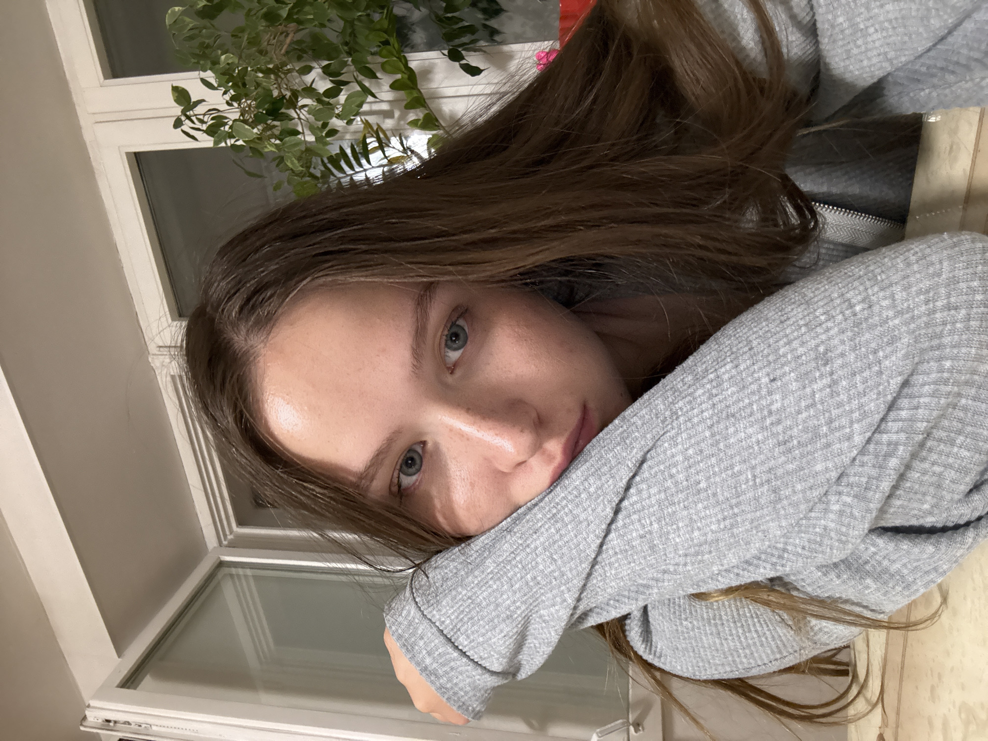 A young woman with long brown hair and blue eyes is resting her face on her arm, which is covered in a light grey, ribbed sweater. A window is behind her, revealing a plant with green leaves. The location is possibly in Chaikovsky, Russia, though there are no specific landmarks to definitively confirm this. The lighting is soft, suggesting an indoor setting. No other people, subjects, objects, actions, or interactions are immediately visible.