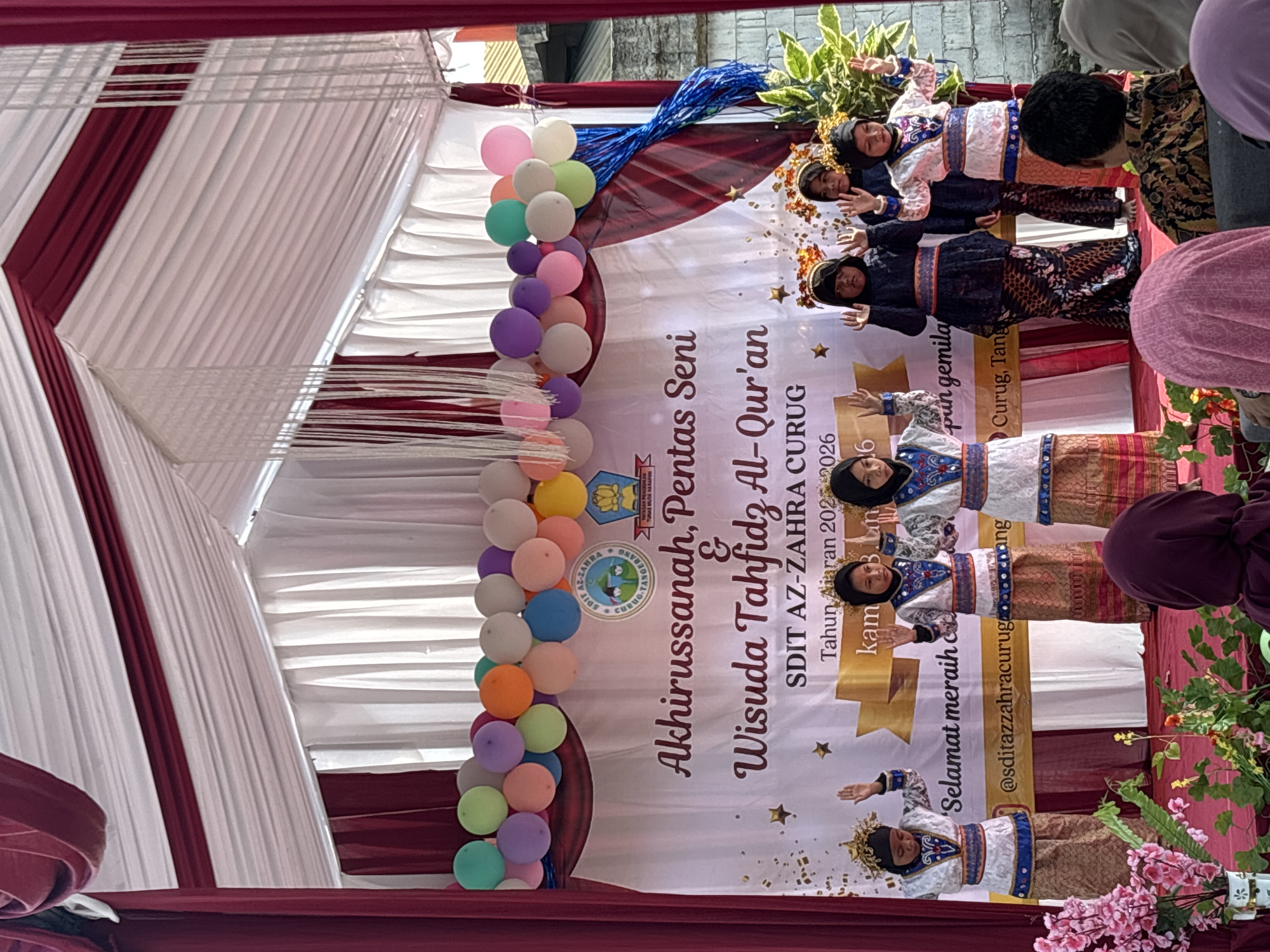 A group of young girls in traditional clothing are performing on a stage in front of a banner that reads "Akhirussanah, Pentas Seni & Wisuda Tahfidz Al-Qur'an SDIT AZ-ZAHRA CURUG Tahun Ajaran 2025-2026". The banner is decorated with balloons and streamers. The girls are wearing colorful outfits with intricate patterns, and some have headpieces. They are all facing the audience with their hands raised. In the foreground, there are flowers and plants. The background shows a tent with red and white fabric. The event appears to be a graduation ceremony or performance for an Islamic school. The location is identified as Sukabakti, Indonesia, based on the banner text which mentions "Curug, Tangerang", a known area within Sukabakti. The audience members are partially visible in the lower part of the image, with one person in a batik shirt in focus.