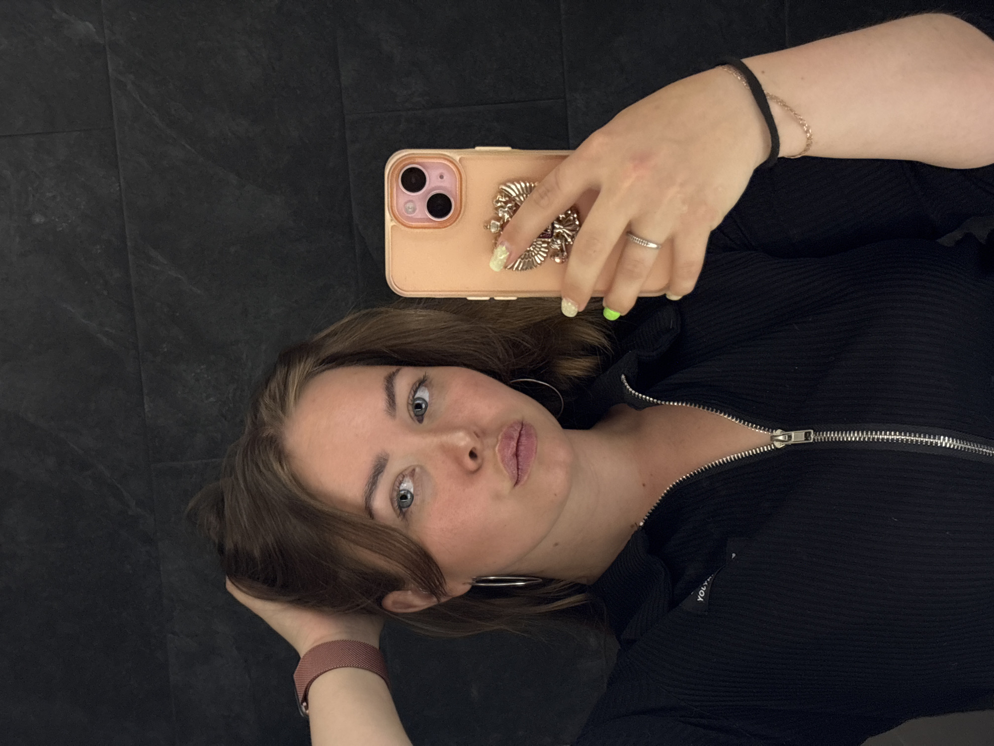A young woman is lying on a dark, tile floor. She is wearing a black, zip-up shirt and large hoop earrings. She is holding a peach-colored cell phone with a decorative grip in her right hand, and her left hand is behind her head. She has a silver ring on her right ring finger and bracelets on her right wrist. She is looking directly at the camera with a slightly pursed expression. The scene is indoors. There is no indication of the city Chaikovsky, Russia.
