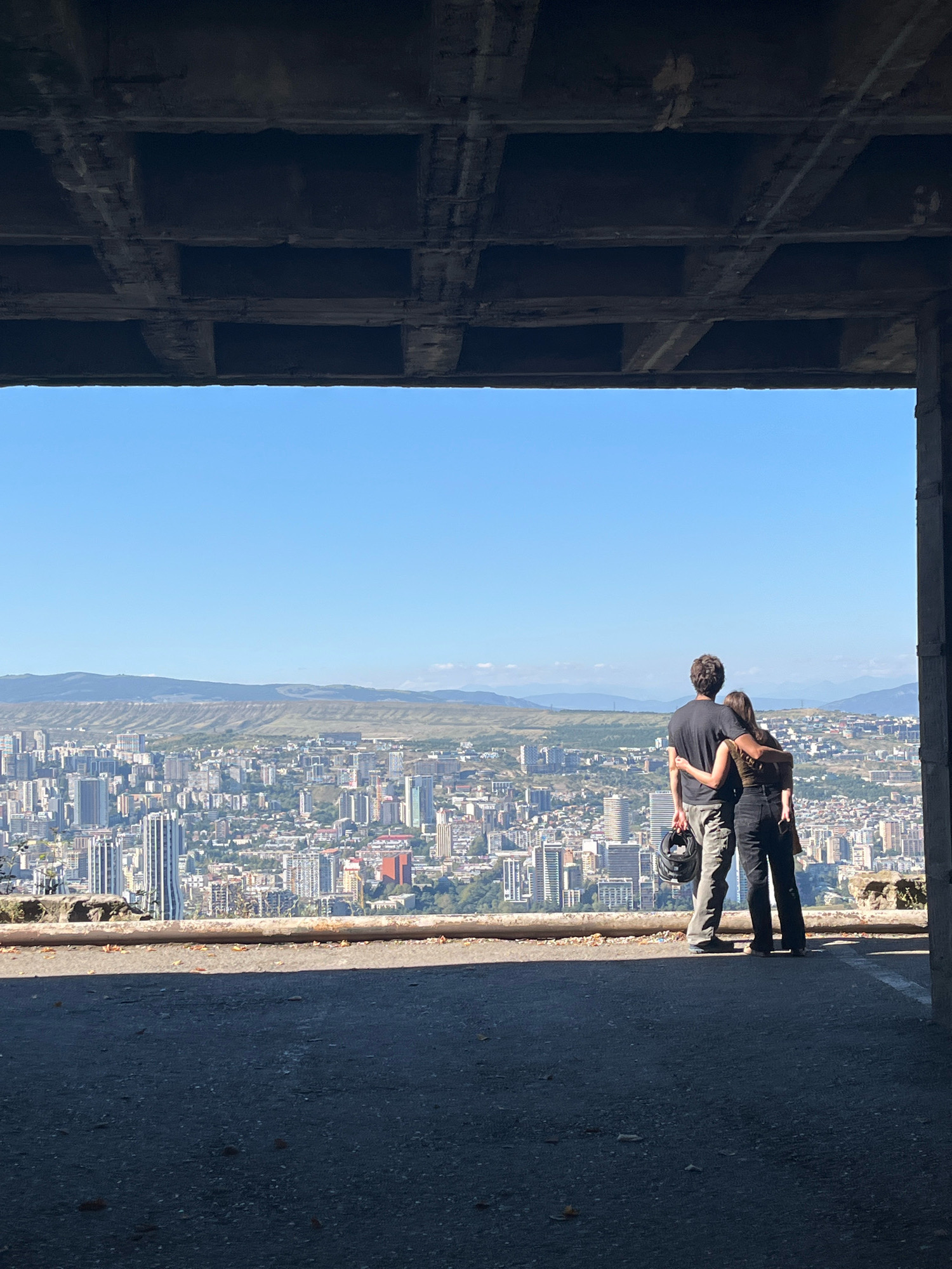 The image shows a couple standing with their arms around each other, looking out over the city of Tbilisi, Georgia. They are framed by a concrete structure, possibly an unfinished building or a viewing platform. The man on the left is wearing a dark t-shirt, cargo pants, and is holding a motorcycle helmet. The woman on the right is wearing a dark top and black pants. The cityscape of Tbilisi stretches out below them, with numerous buildings of varying heights and architectural styles. In the distance, rolling hills and mountains form the backdrop under a clear blue sky. The foreground is a dark, asphalted area, suggesting they are on an elevated vantage point.