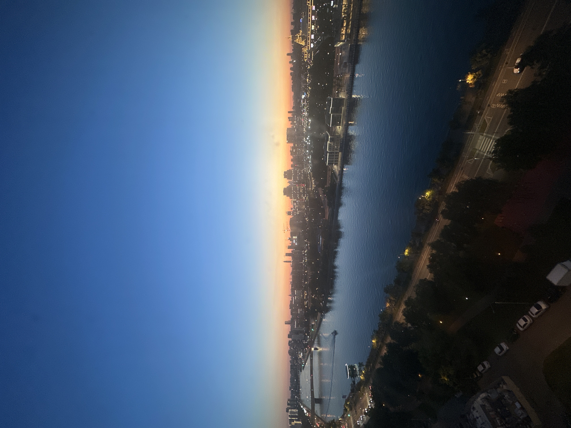 The image shows a panoramic view of Moscow, Russia, at dusk. The sky transitions from a deep blue at the top to hues of orange and pink near the horizon, indicating the setting sun. A wide river, likely the Moskva River, occupies the middle ground, reflecting the city lights.
On the far bank of the river, a cityscape is visible, characterized by numerous illuminated buildings, including skyscrapers and other urban structures. Lights from the city create a warm glow against the darkening sky. A bridge spans the river in the distance, with traffic visible on it. Boats are also present on the water, one of which is leaving a subtle wake.
Along the near bank of the river, a tree-lined promenade or park is visible, with lampposts casting pools of light. Adjacent to this, a road with visible lane markings and streetlights is present, and several cars are parked or driving along it. In the foreground, a dark area with trees and a street with parked cars suggests a residential or urban area. Several white cars and a white utility vehicle are parked on the side of the road.
The overall impression is that of a vibrant city settling into the evening, with the natural beauty of the dusk sky contrasting with the man-made lights of the urban landscape.