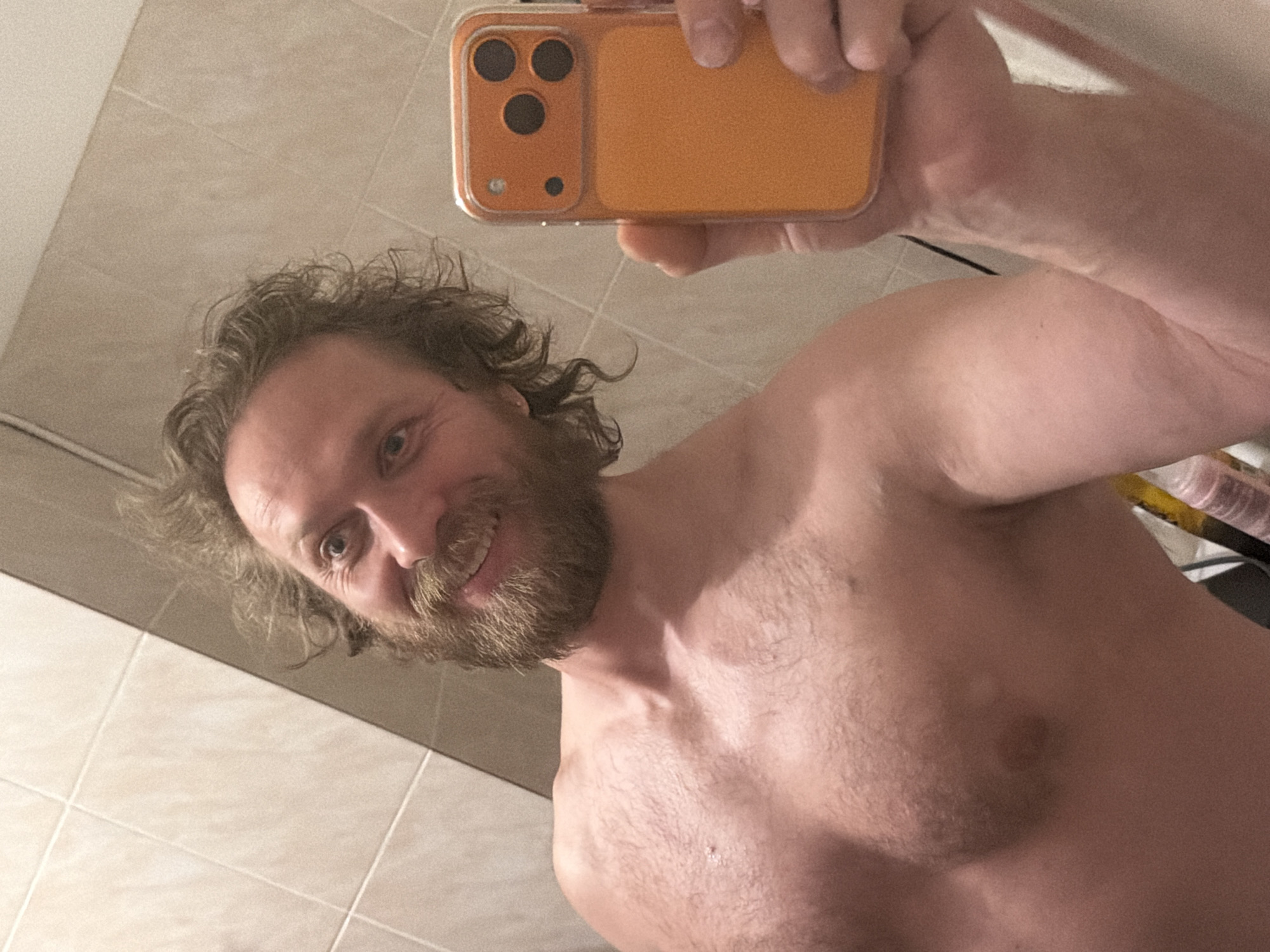 A shirtless man with a beard and wavy hair is taking a selfie with an orange smartphone. The background is a bathroom with beige wall tiles. The location cannot be determined to be in Saint Petersburg, Russia based on the given information.