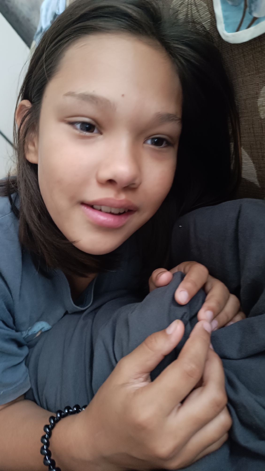 A close-up shot focuses on the face of a young girl lying down. She has dark hair that falls around her face and is wearing a grey t-shirt. Her eyes are open and looking towards the camera, with a slight smile on her lips. Her hands are visible, one of which is holding a dark grey blanket. She is also wearing a black beaded bracelet on her wrist. The background is blurred, with hints of a patterned fabric, possibly a couch or bed, and a light blue patterned object. The lighting is soft, suggesting an indoor setting.