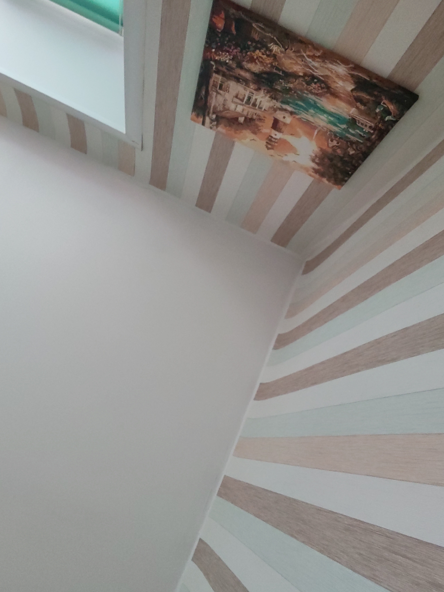 The image shows an interior corner of a room. The walls are decorated with striped wallpaper in alternating shades of light brown and off-white. A painting hangs on the ceiling, depicting a landscape with buildings and water. A window frame is visible in the upper left corner, painted white with a green tint. There are no people visible. The location is unknown, though the prompt mentions the city of Izhevsk, Russia.