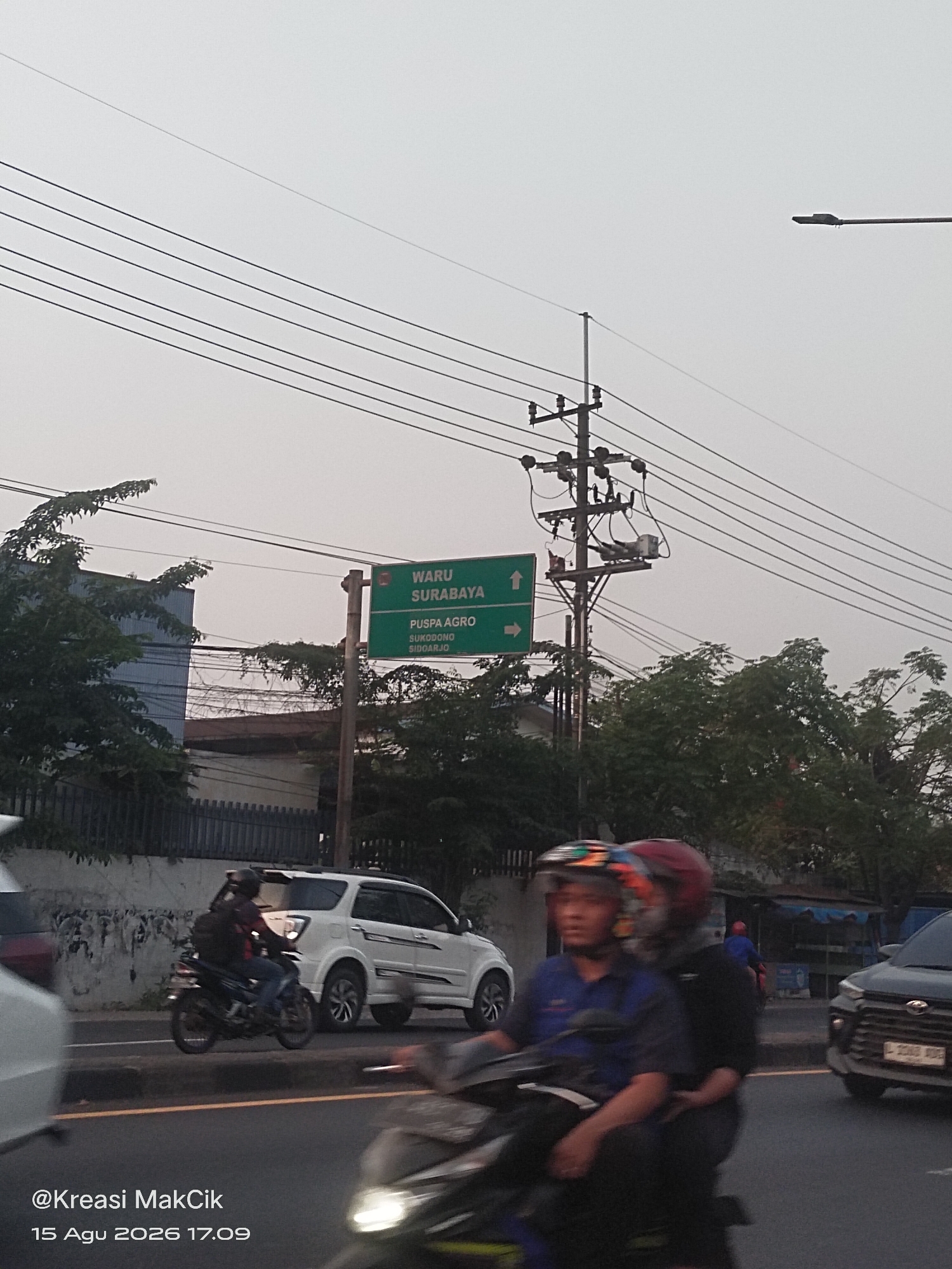 In Kletek, Indonesia, an overcast sky hangs above a street scene. A green directional sign points towards "WARU SURABAYA" straight ahead and "PUSPA AGRO SUKODONO SIDOARJO" to the right. Electrical wires crisscross the sky, supported by a utility pole. On the road, a motorcycle carries two individuals wearing helmets, with the driver in a blue shirt and the passenger in a dark shirt. Behind them, a white SUV is parked. To the right, a black car with a license plate ending in "XDE" is visible. The image is watermarked with "@Kreasi MakCik" and "15 Agu 2026 17.09".