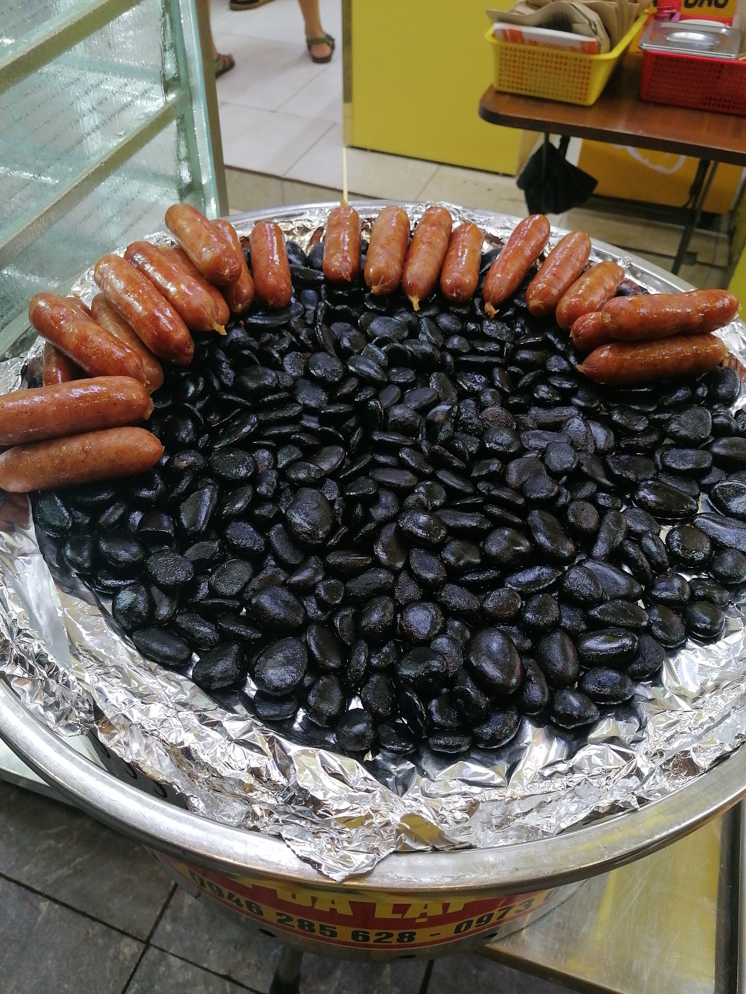 A close-up, eye-level shot depicts a round, metal food vendor's tray lined with crinkled aluminum foil, displaying various items for sale. The tray is largely filled with a dense bed of small, dark, shiny, irregularly shaped objects, resembling roasted or boiled seeds or beans, creating a contrasting texture. Around the rim of this bed, forming an almost complete circle, are numerous cooked sausages. These sausages are plump, light to medium brown, and appear slightly greasy, suggesting they have been grilled, fried, or steamed. Some of the sausages are skewered with thin wooden sticks.
In the background, on the upper left, a frosted or textured glass/plastic partition is visible. To the upper right, part of a simple wooden table holds a yellow plastic basket containing paper bags or similar items, and a red plastic basket next to it. Further in the background, a person's lower legs and feet, clad in dark sandals on a light-colored tiled floor, are visible. The overall setting appears to be a casual outdoor or semi-outdoor food stall, likely within a market or street food area.
Partial text is visible on the side of the metal tray, including the numbers "0946 285 62" and a fragment that might be "LAI," likely indicating a vendor's contact information. No specific time of day or weather conditions are discernible, but the scene suggests commercial activity.