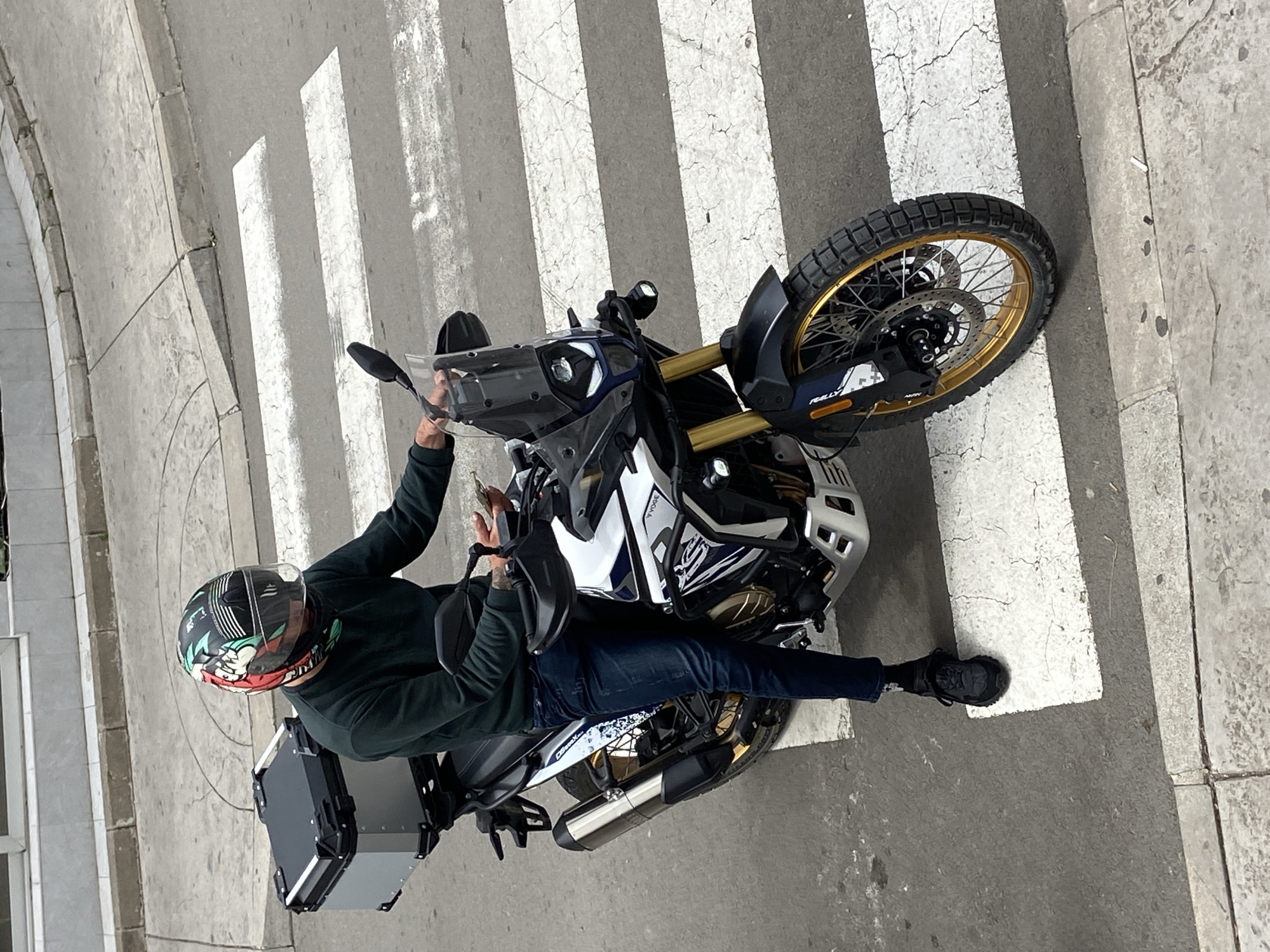 A person wearing a helmet, a dark green long-sleeved shirt, dark jeans, and black shoes is sitting on a motorcycle in a crosswalk. The motorcycle is predominantly white, black, and gold, with what appears to be rally decals. The person is holding onto the handlebars and appears to be looking at something. A black cargo container is mounted on the rear of the motorcycle. The crosswalk is marked with thick white stripes on a grey asphalt surface. The environment shows signs of urban wear and tear, including cracks and discoloration in the pavement. Given the architecture and general urban characteristics, it's possibly located in Budva, Montenegro.