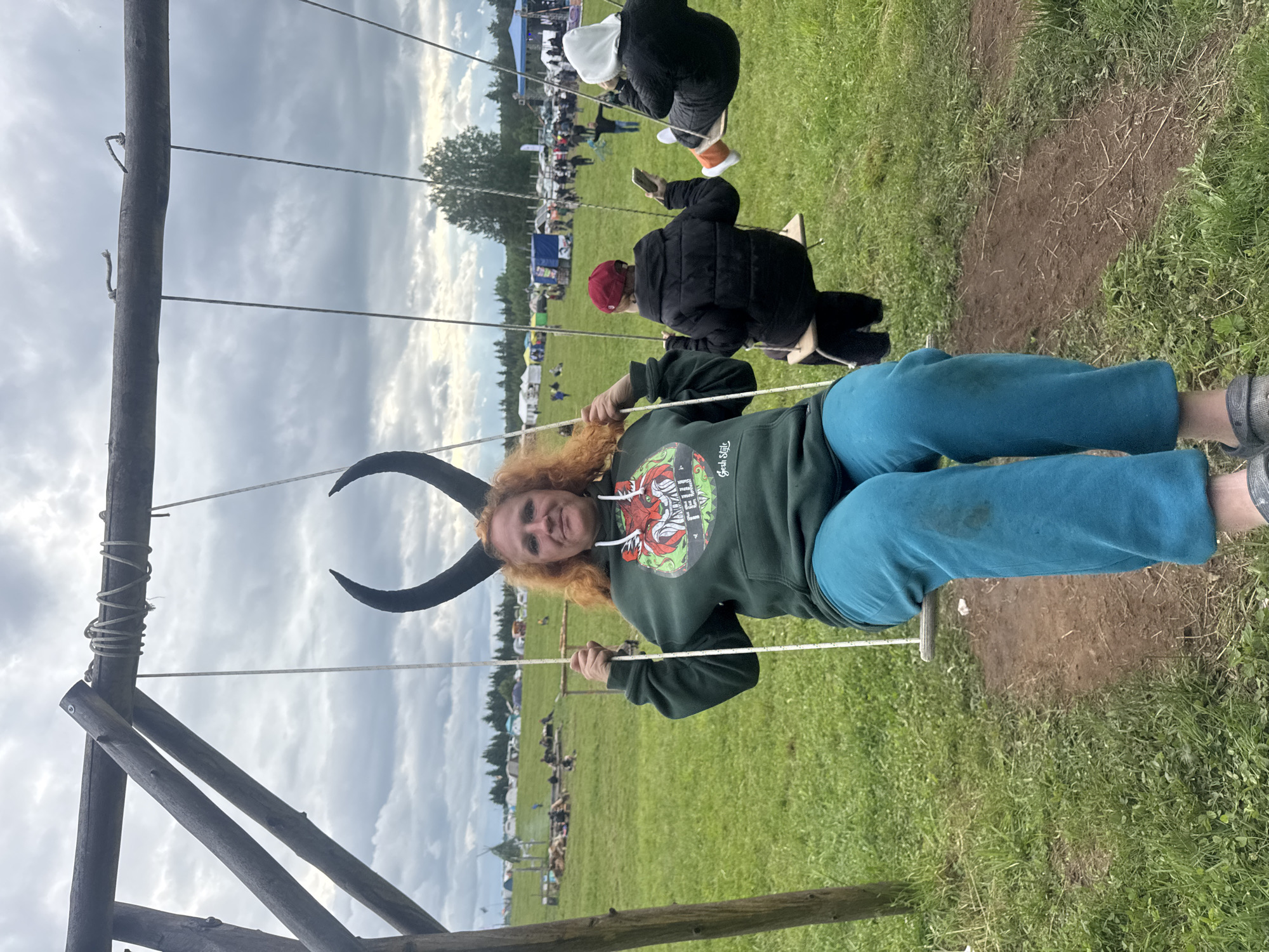 The image depicts an outdoor gathering or festival in a large, open grassy field in Bol'shaya Kivara, Russia, under an overcast sky. A rustic wooden swing set, constructed from logs, occupies the foreground.
An adult female with wavy, shoulder-length red hair and large, black, upward-curving horns on her head is centered on the leftmost swing. She wears a dark green hooded sweatshirt featuring a circular graphic with a red, stylized animal (resembling a fox or wolf) and the text "ГЕШ" and "Gesh Style". Her lower body is clad in bright blue sweatpants, with a dark smudge visible on the right thigh, and she wears grey clogs or sandals. She holds the swing ropes with both hands and looks directly at the viewer with a slight smile.
To her right, a second individual sits on an adjacent swing, their back partially obscured by the first person. This individual wears a dark red cap and a dark (likely black) puffer jacket, holding a light-colored smartphone in their right hand. Further to the right, a third person is seated on another swing, facing away from the camera. This individual wears a white hood over their head, a dark puffer jacket, and bright orange socks with light-colored shoes visible on their right foot.
The ground beneath the swings consists of worn earth mixed with patches of green grass. In the mid-ground and background, an expansive green field stretches towards a distant treeline. Numerous tents of various sizes and colors are scattered across the field, indicative of a large temporary camp or market. In the far left background, a group of people is gathered around what appears to be a fire pit with log seating, near a small, conical teepee-like structure. In the far right mid-ground, a larger blue canopy or stage structure is visible, with more distant figures and some vehicles present. The overall lighting is diffused due to the heavy cloud cover.