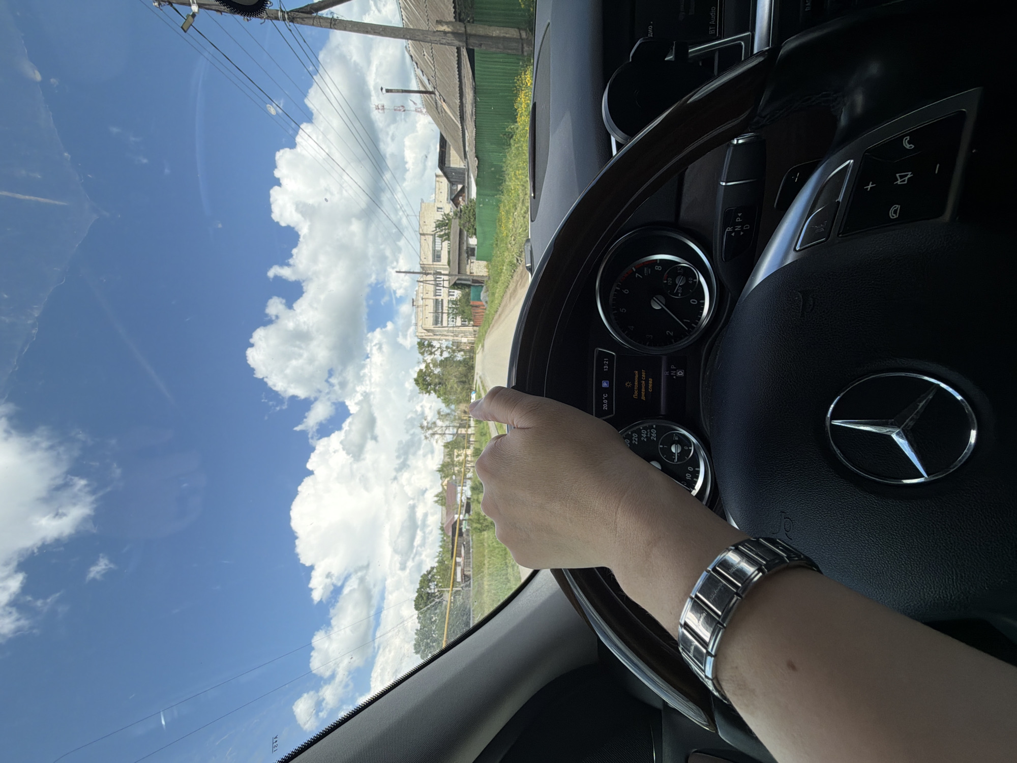 A first-person perspective shot from inside a Mercedes-Benz car shows a person's left hand on the steering wheel, wearing a silver link watch. The car is driving down a paved road with green grass and some yellow flowers on the sides. In the background, there are several houses and buildings, some with green fences. Tall power lines and a telecommunications tower are visible on the right side of the road. The sky is blue with scattered white clouds. The car's dashboard displays the time as 13:21, the outside temperature as 20.0°C, and a gear indicator showing "P" for park. The car is likely in a rural or semi-urban area in Mezhevoy, Russia, given the architectural style of the buildings and the presence of power lines.