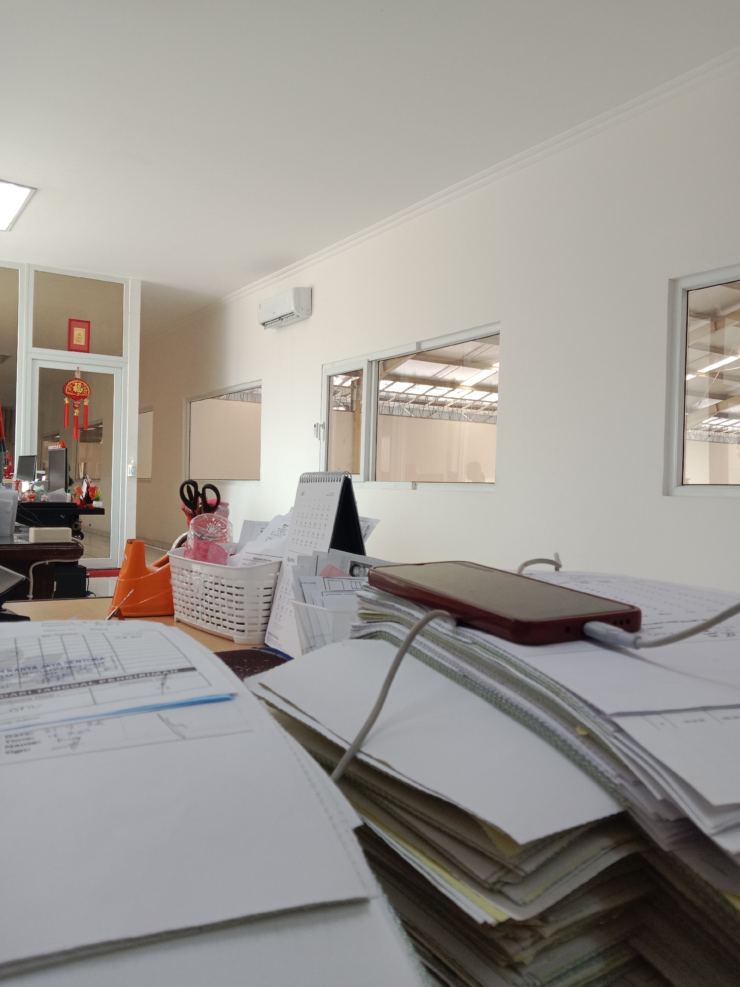 This is a medium shot of an office desk with papers, a phone, and office supplies. The room appears to be an office with a window looking out onto an industrial or warehouse-like structure. The ceiling is white with subtle molding and a fluorescent light fixture on the left. An air conditioning unit is mounted on the wall. On the desk, there is a stack of papers, some of which are covered in writing and grids. A red-cased smartphone is plugged in and lying on top of some documents. To the left of the phone is a white basket containing scissors and some pink items. An orange desk organizer is also visible. Chinese New Year decorations, including red lanterns and a framed red character, are hanging in the doorway of an adjacent room. The overall scene suggests an active workspace, possibly in an industrial company located in Semarang, Indonesia, given the decorative elements.