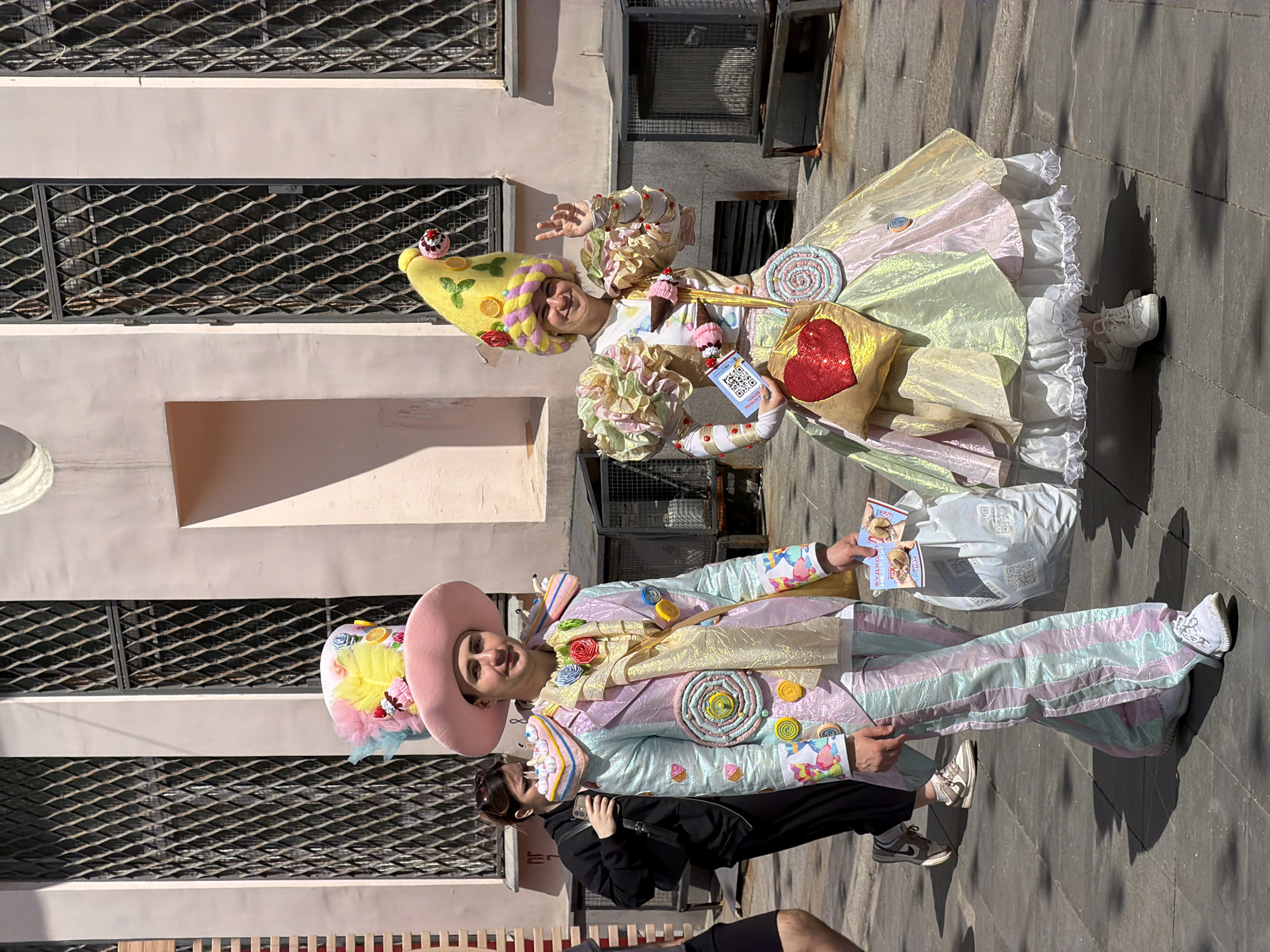 The image, oriented vertically and rotated 90 degrees to the right, captures two individuals in elaborate, candy-themed costumes posing on a sunlit city street in Moscow, Russia.
The figure on the left, a man, wears a pastel-striped suit in light blue, pink, and yellow, adorned with numerous colorful faux sweets like lollipops, cupcakes, and rosettes. He sports a large, soft pink hat resembling a cake, topped with fluffy decorations and miniature cakes. He is smiling and holds two small brochures featuring images of cakes and the Russian text "БУДЬ ВКУСНЫМ" (Be Tasty/Delicious).
To his right, a woman is dressed in a voluminous, iridescent gown in pastel shades of pink, yellow, and green, layered over a white ruffled underskirt. Her outfit includes large, decorative sleeve puffs and is embellished with similar candy motifs, including a prominent lollipop design on her skirt. She wears a pointed yellow hat, possibly inspired by a lemon meringue pie or cupcake, also topped with a small cake. She smiles, holding a gold bag with a large red sequined heart and a small card displaying a QR code and partial text that appears to start with "E VUDH".
A third person, dressed in a black jacket and pants, is partially visible between the two costumed figures, and legs of a fourth person are seen at the bottom left. The setting is an outdoor urban environment with light-colored buildings featuring dark grates over windows. The bright sunlight casts distinct shadows, indicating a clear, sunny day. Both costumed individuals appear cheerful and engaging, likely participating in a street performance, festival, or promotional event related to the candy theme.