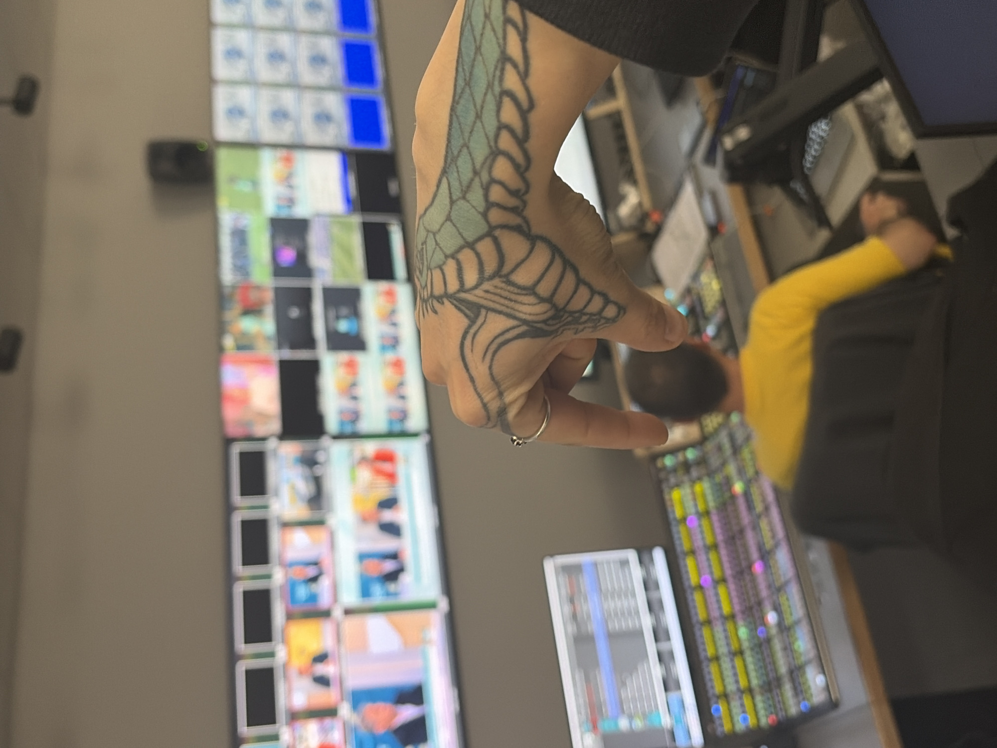 A person with a snake tattoo on their hand is in a room with multiple monitors displaying various images and data. The room appears to be a control room or production studio, based on the equipment and the presence of another individual in a yellow shirt in the background. The setting suggests a professional environment. Location cannot be determined without further information.