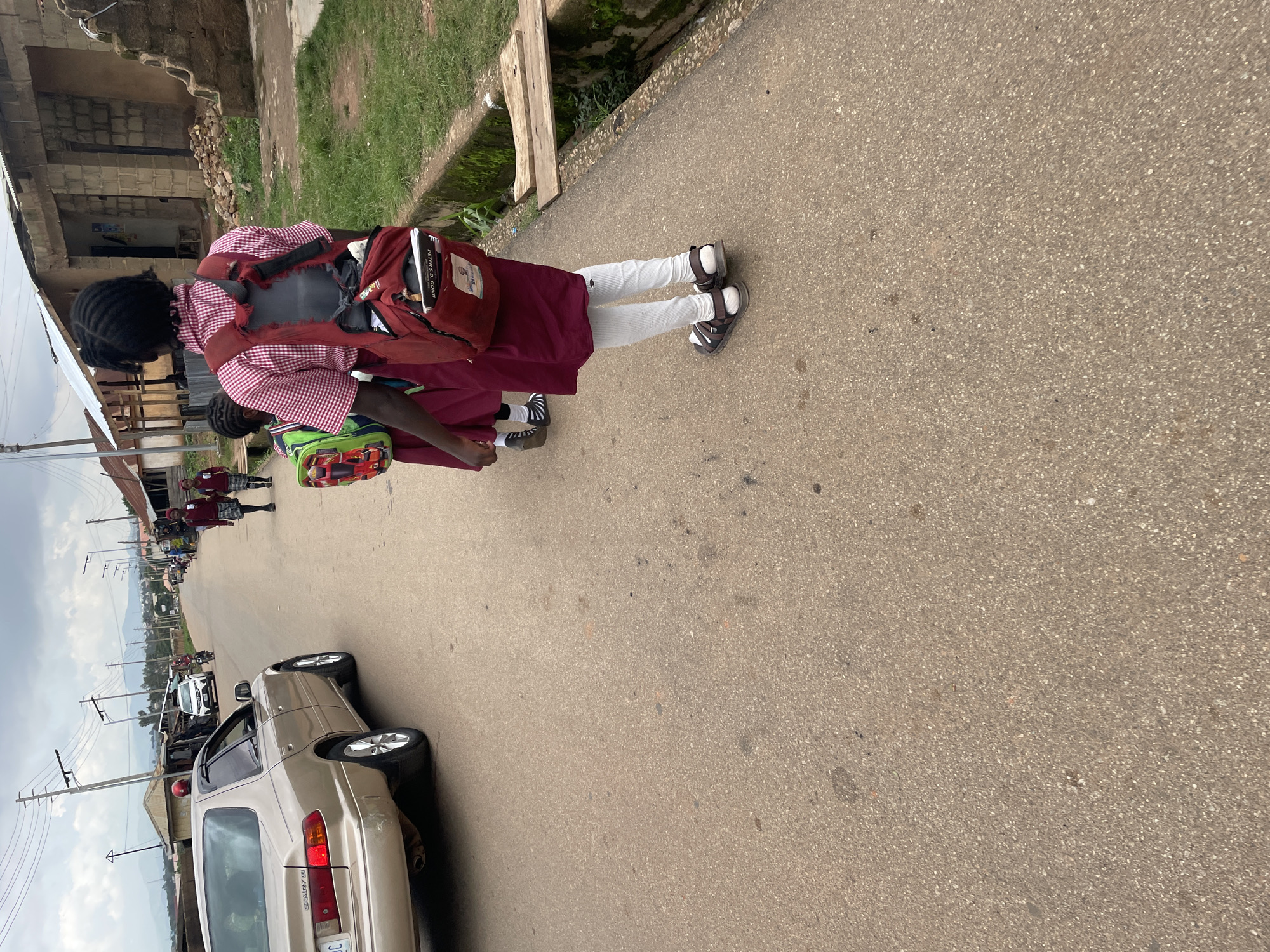 In Jos, Nigeria, a golden Toyota Camry is parked on the side of a paved road. Two young girls, presumably students, walk away from the camera on the same road. They are wearing matching maroon skirts and white socks, and one girl has a red and white checkered shirt. Both are carrying backpacks. The girl in the foreground has a red backpack with the words "PETER S. GONII" visible. The other girl carries a green backpack with a cartoon car design. Buildings, some appearing unfinished, line the right side of the road, along with a drainage ditch. A second white car and a motorcycle are visible further down the road, as well as other pedestrians. The sky is overcast.