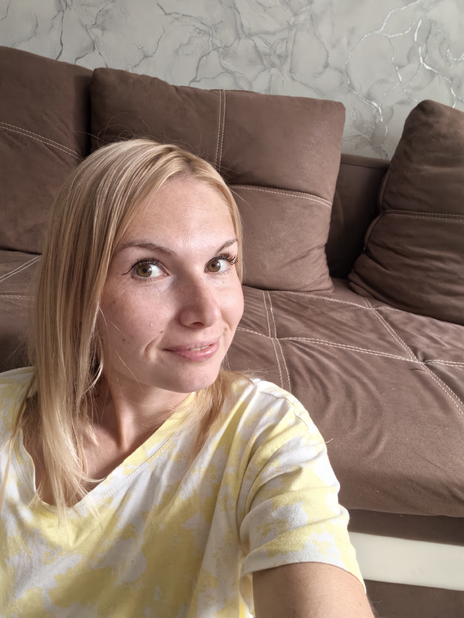 A Caucasian woman with blonde hair is taking a selfie in a living room. She is wearing a yellow and white tie-dye t-shirt. Behind her is a brown couch with textured pillows, and a wall with a marble-like pattern. The scene is lit by natural light. The image does not provide any information about the city of Sterlitamak, Russia.
