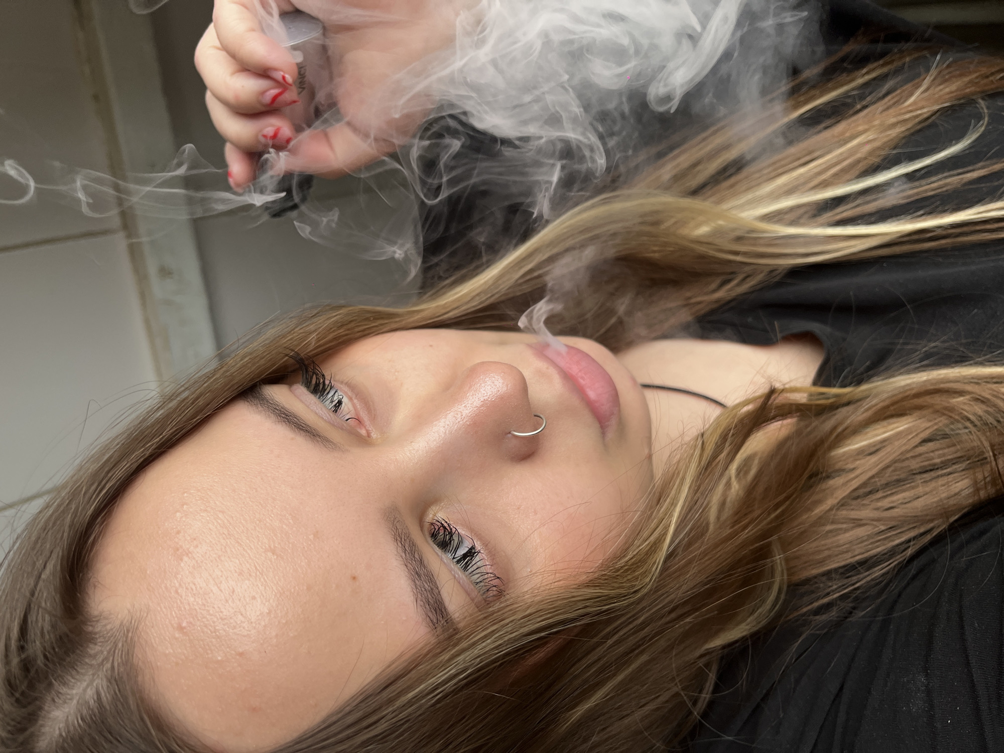 A young woman with light brown and blonde hair lies down, exhaling vapor. She has a nose ring on the left side. Her left hand holds a vape device emitting vapor. She is wearing a black shirt. The background appears to be a light-colored wall. It is impossible to determine if the picture was taken in Novopokrovskaya, Russia.